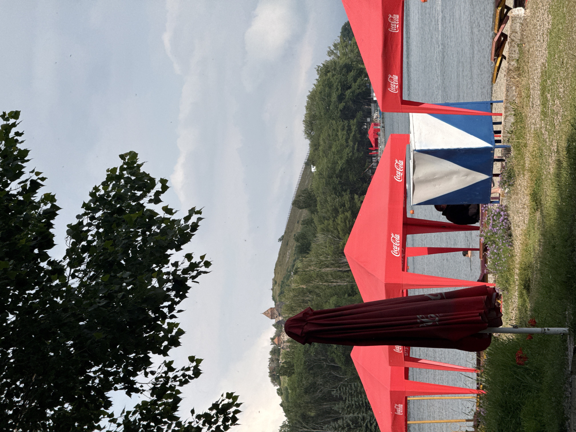 The image depicts a waterfront scene in Sevan, Armenia, likely during a warm season. In the foreground, several red canopies with Coca-Cola branding are set up, suggesting a vendor or relaxation area. A closed red umbrella stands prominently in front of the canopies. The ground is covered with grass and some scattered flowers. Beyond the canopies, a calm body of water is visible, with a few people in the distance. In the background, a lush green hillside rises, topped with a historical building complex with domed structures. The sky is overcast with scattered clouds. The overall atmosphere suggests a leisure or tourist location.