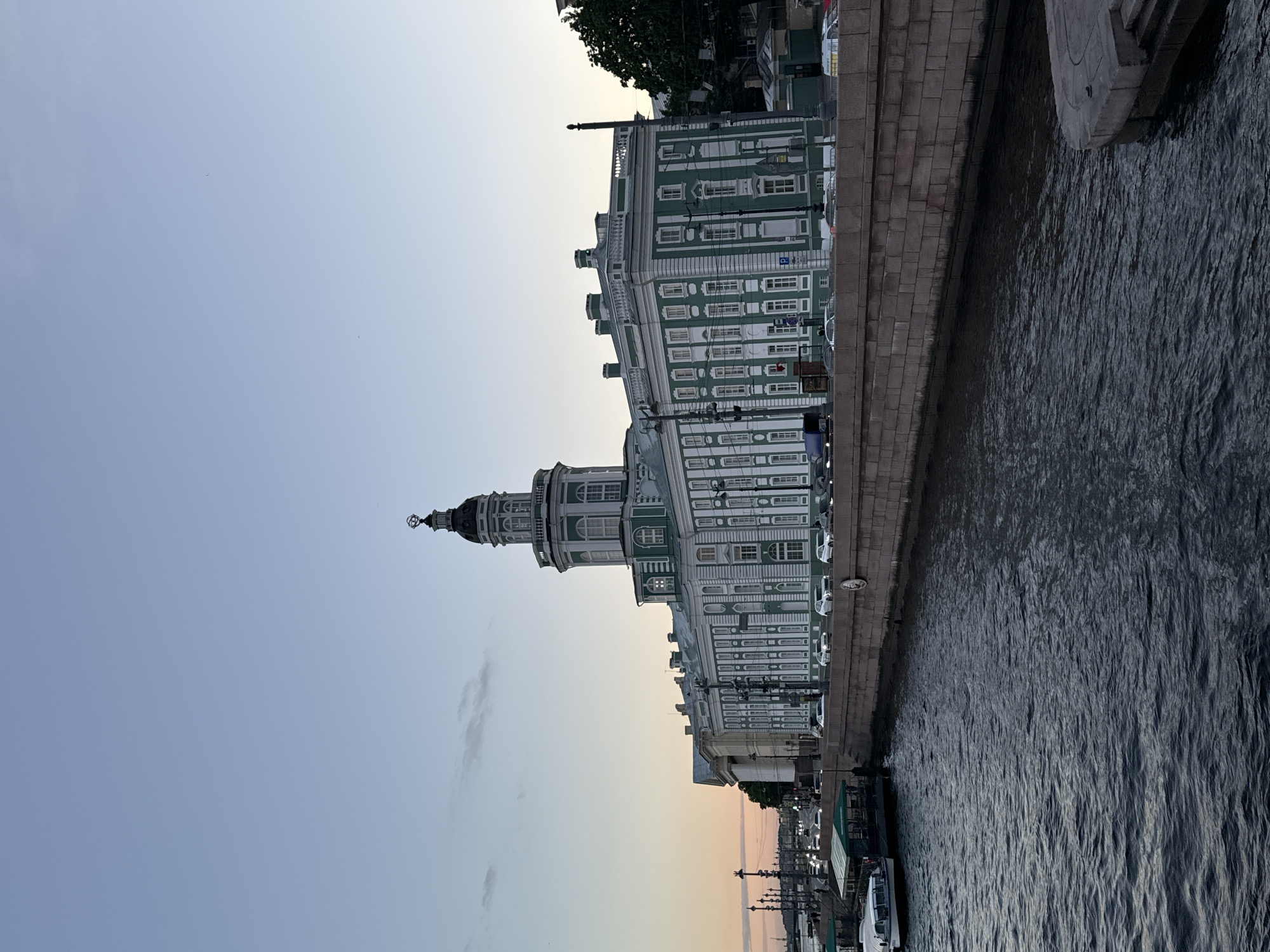 The image shows the Kunstkamera museum in Saint Petersburg, Russia, viewed from across the Neva River. The sky is a clear, pale blue, transitioning to a pale orange near the horizon, indicating it is either sunrise or sunset. The Kunstkamera building is large, white, and green, with a distinctive domed tower. It stands on a stone embankment, with cars parked along the street in front of it. On the left side of the image, a body of dark, rippling water, presumably the Neva River, occupies the foreground. A boat with a green awning is docked on the riverbank. The overall scene is calm and serene, showcasing the architecture of the historic museum against a tranquil sky.