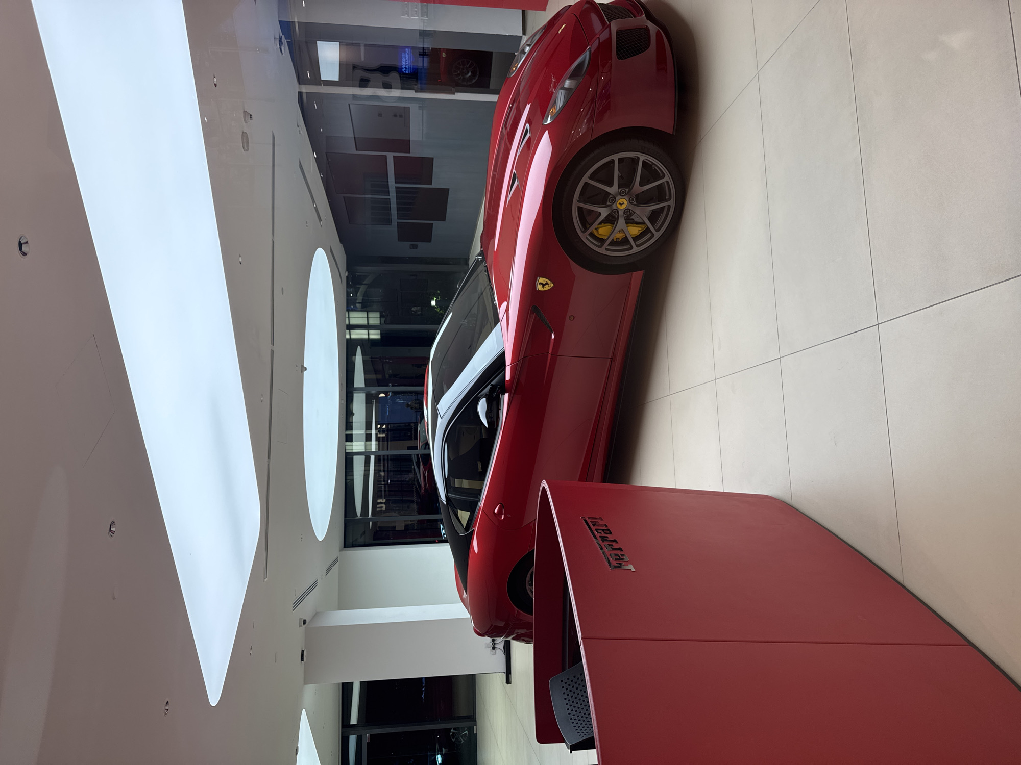 A red Ferrari sports car is displayed indoors, likely in a showroom or dealership. The car is positioned near a red stand with the Ferrari logo on it. The interior of the building features white walls and a modern lighting design with curved, illuminated panels. A tiled floor is visible. The location appears to be in Kuala Lumpur, Malaysia, but is not possible to confirm. No people are visible in the image.