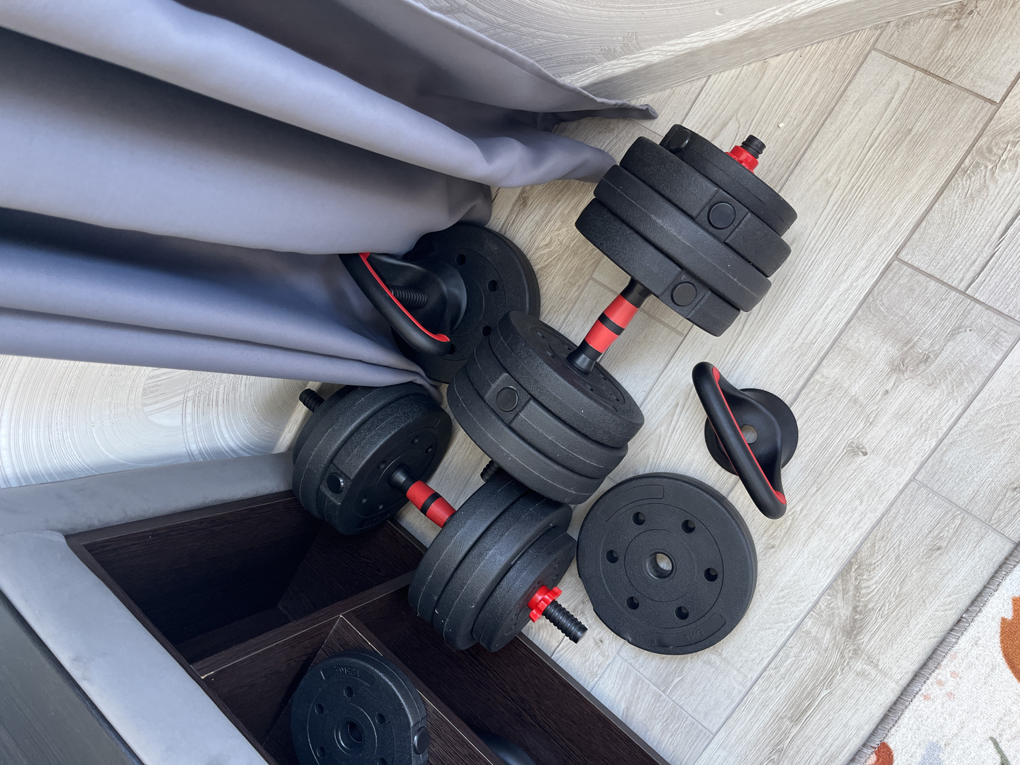 This image captures an indoor scene, likely within a home in Sochi, Russia, showcasing exercise equipment on a light-colored, wood-look laminate floor. The main subjects are a set of black adjustable dumbbells with red accents on their handles and locking mechanisms.
Two fully assembled dumbbells are visible, one prominently in the foreground and another further back, partially obscured by a grey fabric, possibly a curtain or sofa upholstery. Each dumbbell features multiple circular black weight plates attached to a central bar. Several loose black weight plates are also scattered on the floor; some clearly display "3KG" in white text, indicating their weight, and "ONE" is also visible on one plate. Additionally, a black kettlebell-style handle with a red accent is present on the floor, suggesting versatility in the exercise set.
The equipment rests beside a dark brown wooden shelving unit or piece of furniture. A soft grey fabric, possibly a curtain, drapes down the left side of the frame. In the bottom right corner, a small section of a patterned rug with white, orange, and floral elements is visible, adding a touch of domesticity to the scene.
No specific activity is taking place; the equipment appears to be stored or temporarily laid out. The bright, even lighting suggests it is daytime. The overall impression is that of a home fitness setup.