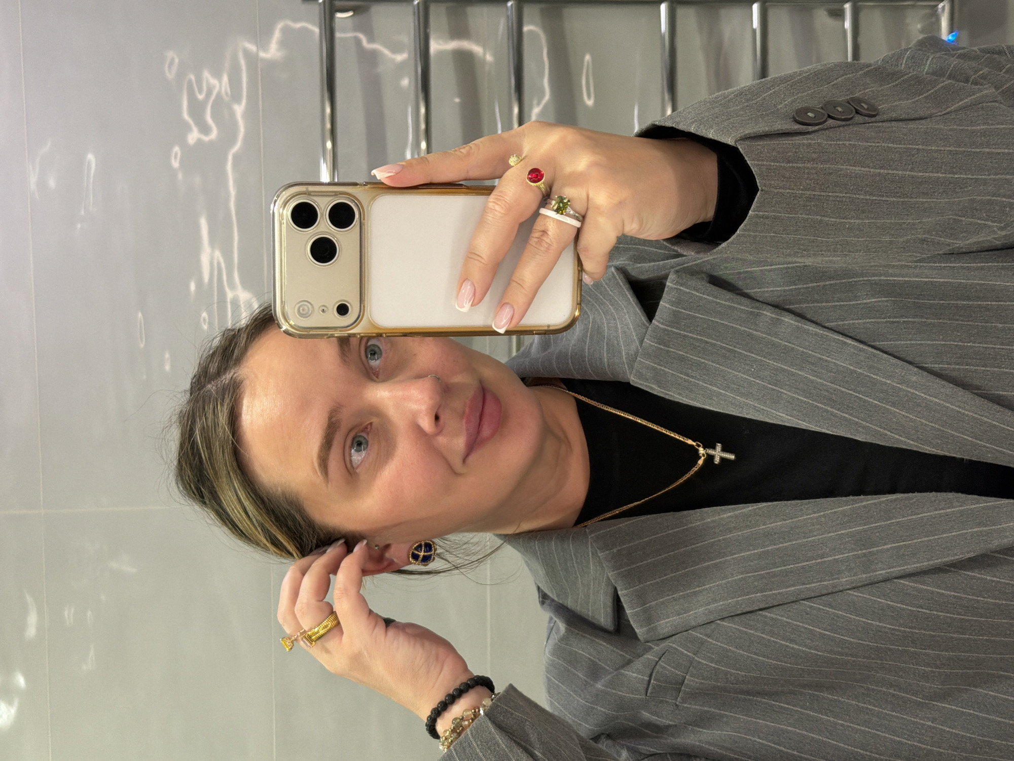 A woman takes a selfie in a room with a gray and white wall and chrome towel racks. She holds an iPhone with a gold and white case. She wears a gray pinstripe blazer over a black shirt and a gold necklace with a cross pendant. She has gold rings on both hands, beaded bracelets, and blue and gold earrings. There is no indication of this image being in Khimki, Russia.
