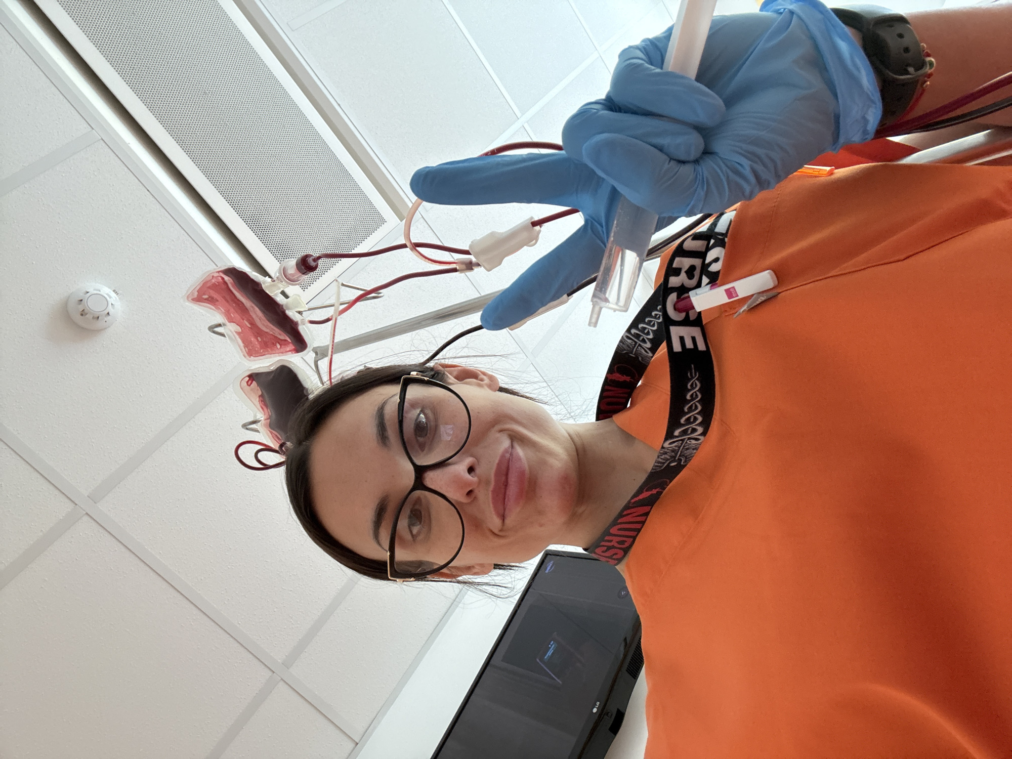 A young woman wearing glasses and an orange scrub top poses for a selfie, indoors. She is wearing blue gloves and making a peace sign with her gloved hand. There is medical equipment, including IV tubes and blood bags, in the background attached to an IV pole, suggesting a medical setting. Her lanyard indicates she is a nurse. A computer monitor is visible. The ceiling is white with square tiles, a light fixture, and a fire alarm. There is no information to confirm this photo was taken in Pleven, Bulgaria.