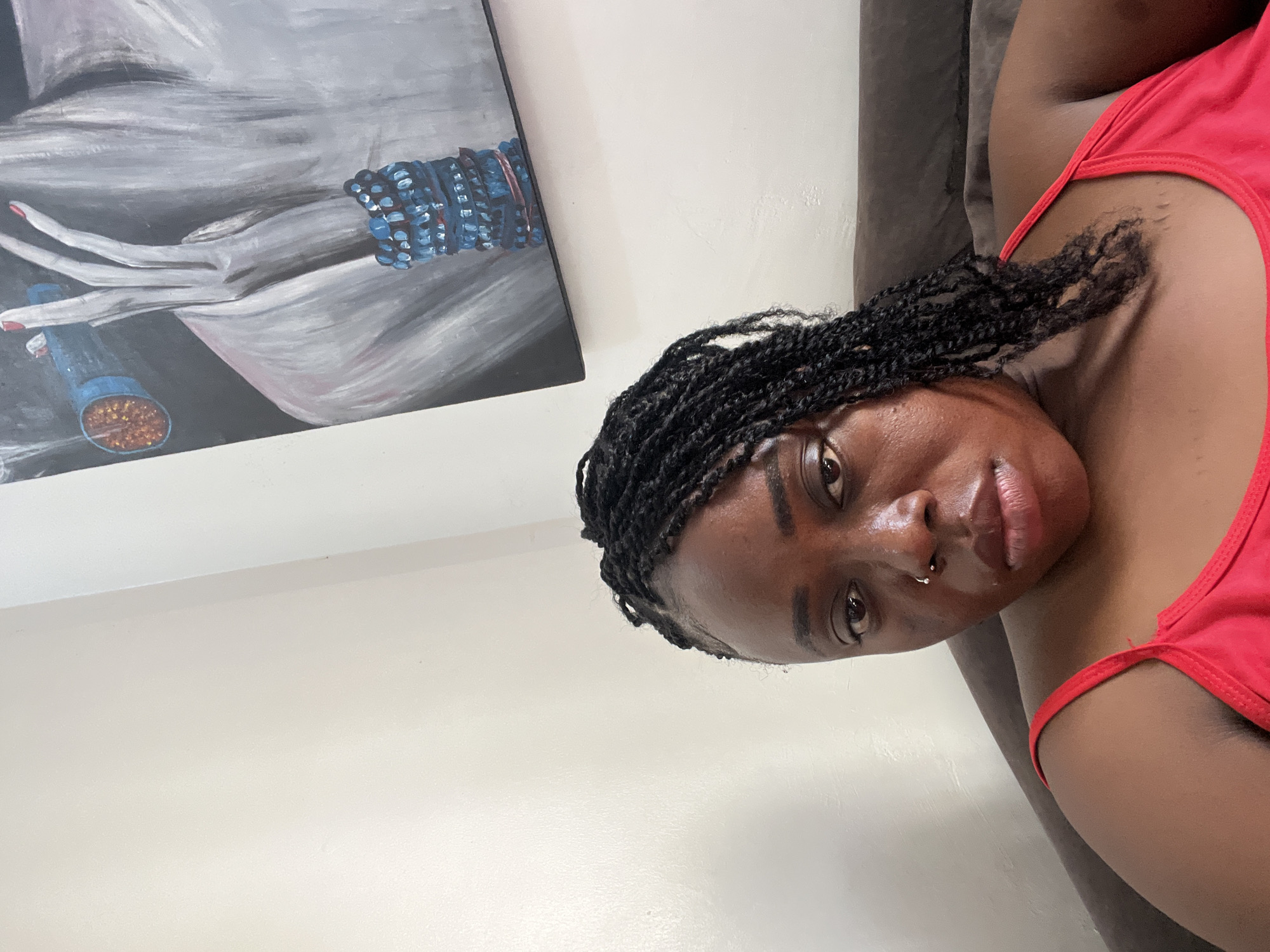 A woman with braided hair and a red top is sitting on a brown couch in front of a white wall with a painting. The painting depicts a close-up of a person's torso and hand with a blue bracelet. The woman is looking at the camera with a neutral expression and has a nose ring. The lighting is natural and appears to be coming from the side. There are no specific landmarks or contextual clues to place this image in Abuja, Nigeria.
