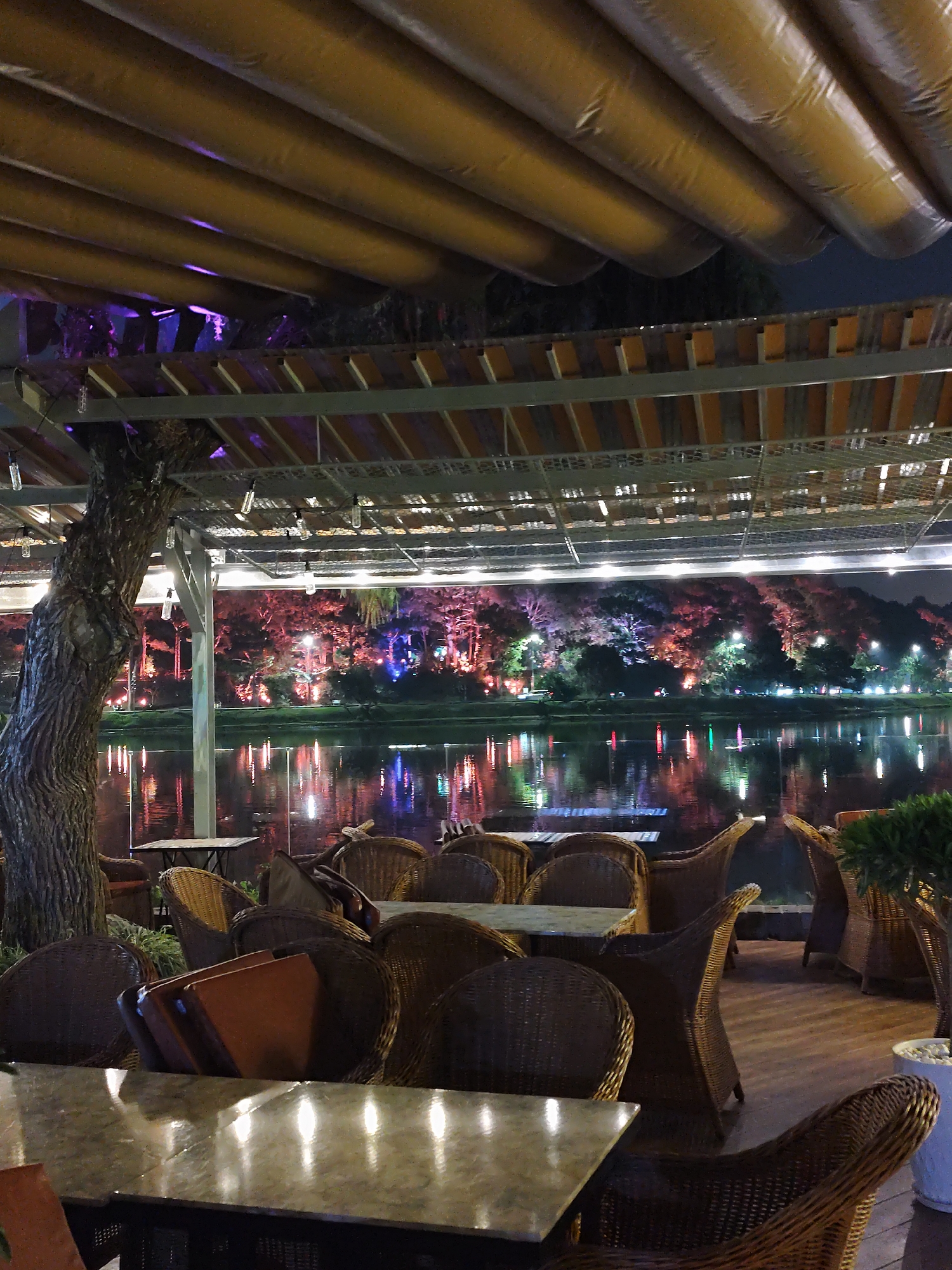 The image captures an outdoor dining or cafe terrace situated on the shore of Xuan Huong Lake in Da Lat, Vietnam, at night. The foreground features an array of unoccupied tables with light-colored tops and numerous dark brown woven wicker chairs; some chairs are stacked. A large, thick tree trunk with textured bark serves as a structural support on the left. Overhead, a beige or tan retractable canopy, composed of parallel padded sections, covers the seating area. String lights with clear bulbs are suspended from a metal grid structure supporting the canopy. Beyond the terrace, the dark surface of Xuan Huong Lake reflects a multitude of colorful lights emanating from the opposite bank. The distant shoreline is lined with trees illuminated in vibrant red, purple, green, and white hues, interspersed with additional streetlights. The sky above is dark, indicative of nighttime. No people are visible within the scene.
