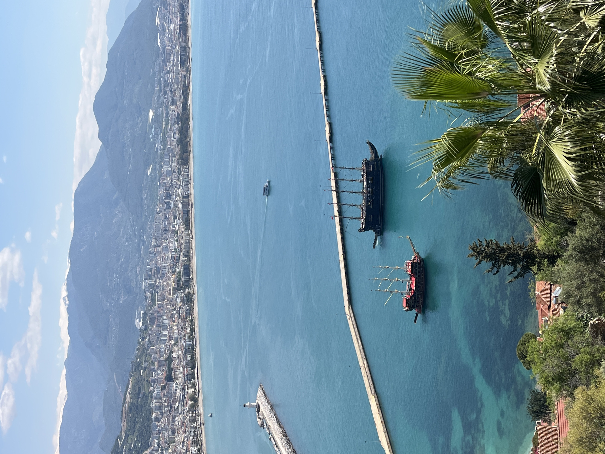 The image shows an aerial view of the coastline of Alanya, Turkey. In the background, there is a mountain range with the city built along its base and the coast. A long concrete pier stretches out into the turquoise sea, with two large pirate-themed ships docked alongside. One ship is primarily black, while the other has red and black features. A smaller boat is visible between the city coastline and the first docked ship. The foreground is partially obscured by a palm tree and other greenery, including a view of some tiled rooftops. The weather appears sunny and clear.