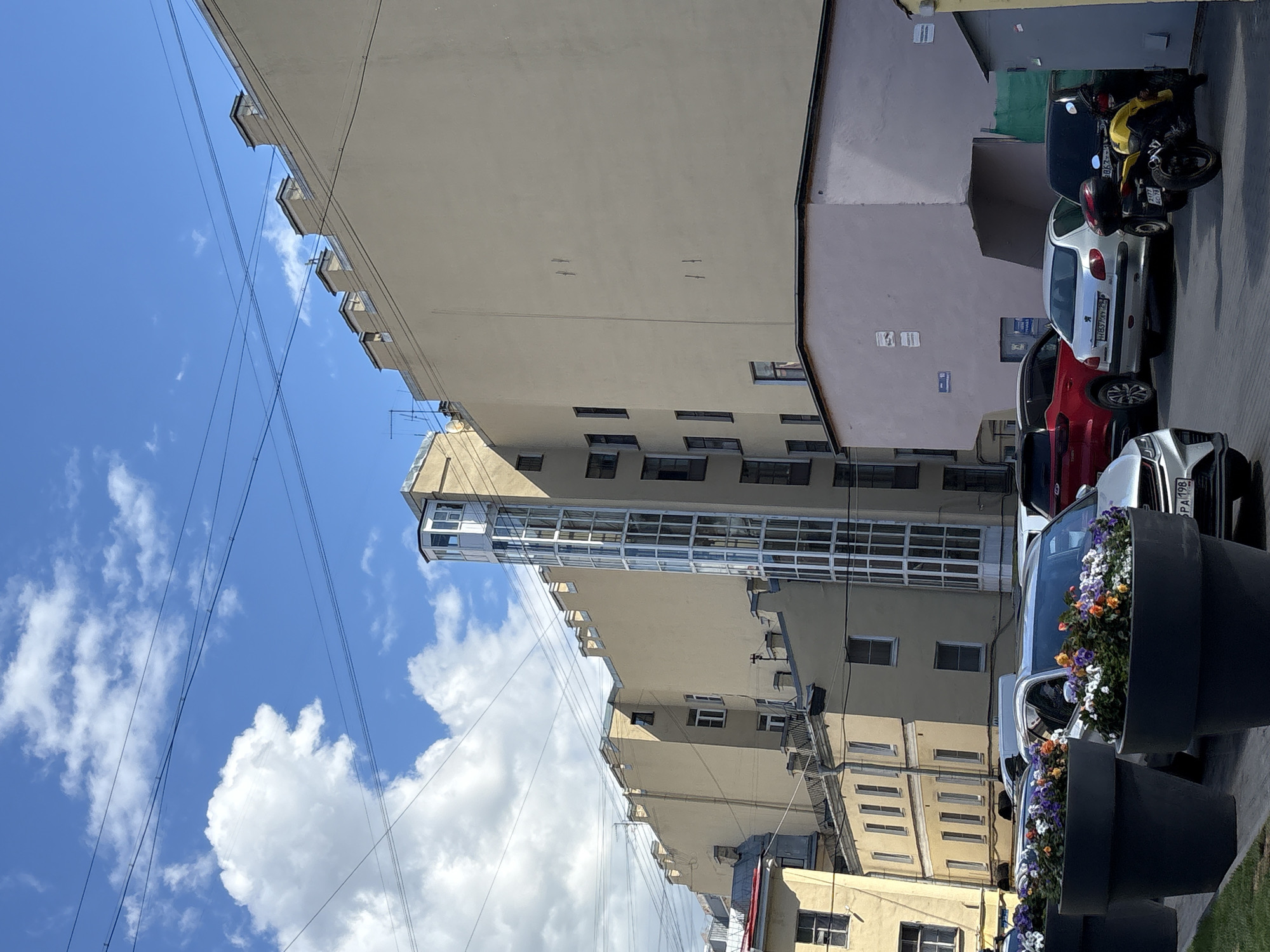 This is an eye-level, wide-angle, exterior shot of a street scene in Saint Petersburg, Russia. The image is oriented sideways. A light-yellow, multi-story building with many windows and balconies dominates the left side of the frame. Adjacent to it, a lighter-colored building with a pinkish hue features a modern, glass-enclosed elevator shaft extending vertically. Further down the street, several parked cars, including a white hatchback, a red sedan, and a silver SUV, are visible. A yellow and black motorcycle is parked behind the white hatchback. Large, dark planters filled with colorful flowers, including white, purple, and orange blooms, are placed in the foreground, obscuring parts of the parked cars. The sky is a bright blue with scattered white clouds, suggesting it is daytime and fair weather. Power lines crisscross the sky. Some visible text on the license plate of the white car reads "H H657MY 198".
