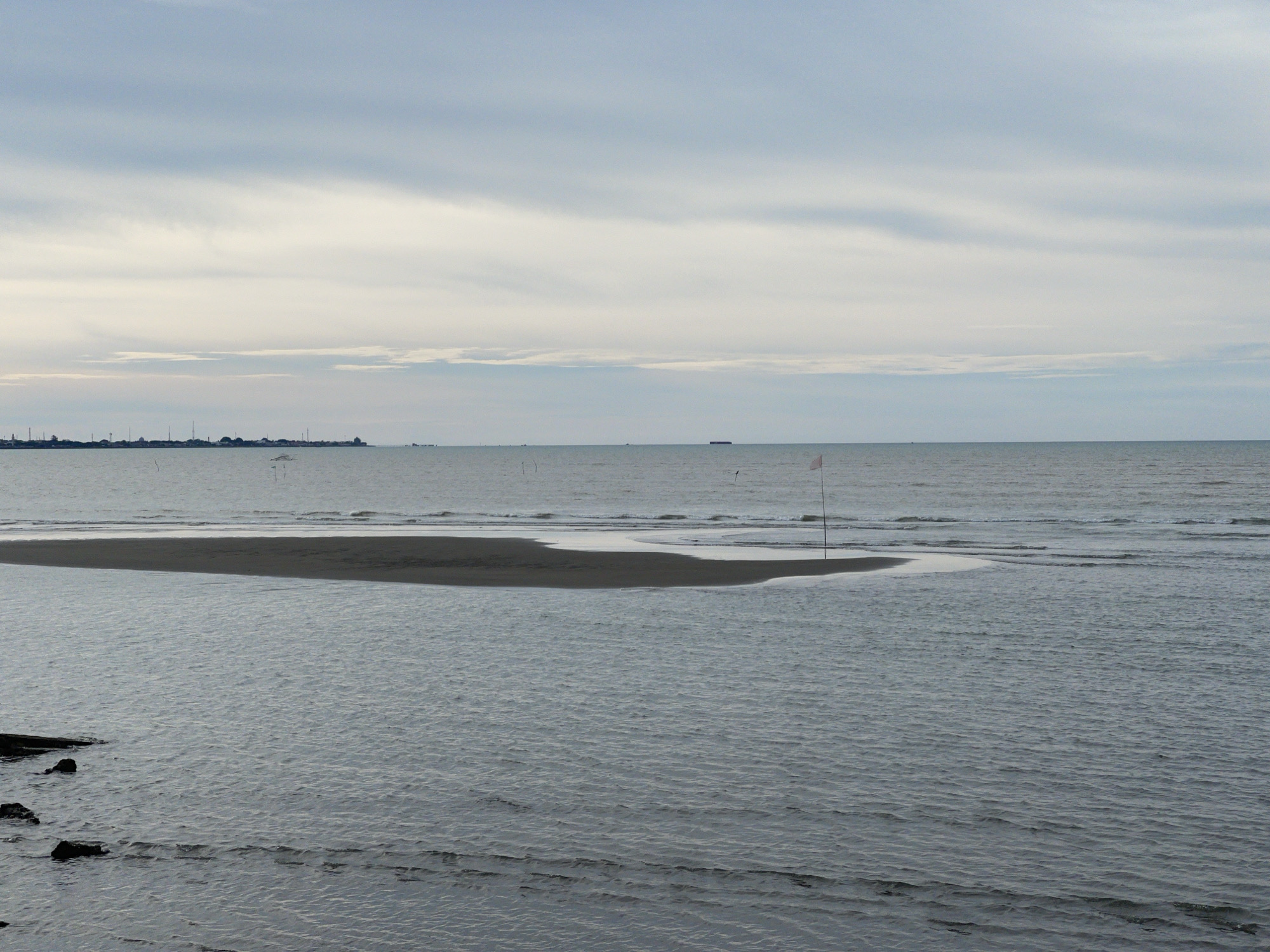 The image depicts a coastal scene in Lancok, Indonesia, likely during overcast weather due to the diffused light and muted colors. The foreground is dominated by shallow, rippling water with dark rocks visible on the left edge. A sandbar, with a darker, wet section and a lighter, drier section, emerges from the water in the middle ground. A single white flag on a pole stands on the edge of the sandbar. The horizon separates the sea from a sky filled with layered, pale gray clouds. In the far distance, a faint, dark line suggests a city or industrial area with numerous vertical structures, possibly including antennas or smokestacks, and a few ships are visible on the water. The overall impression is one of a quiet, possibly low-tide, coastal environment.