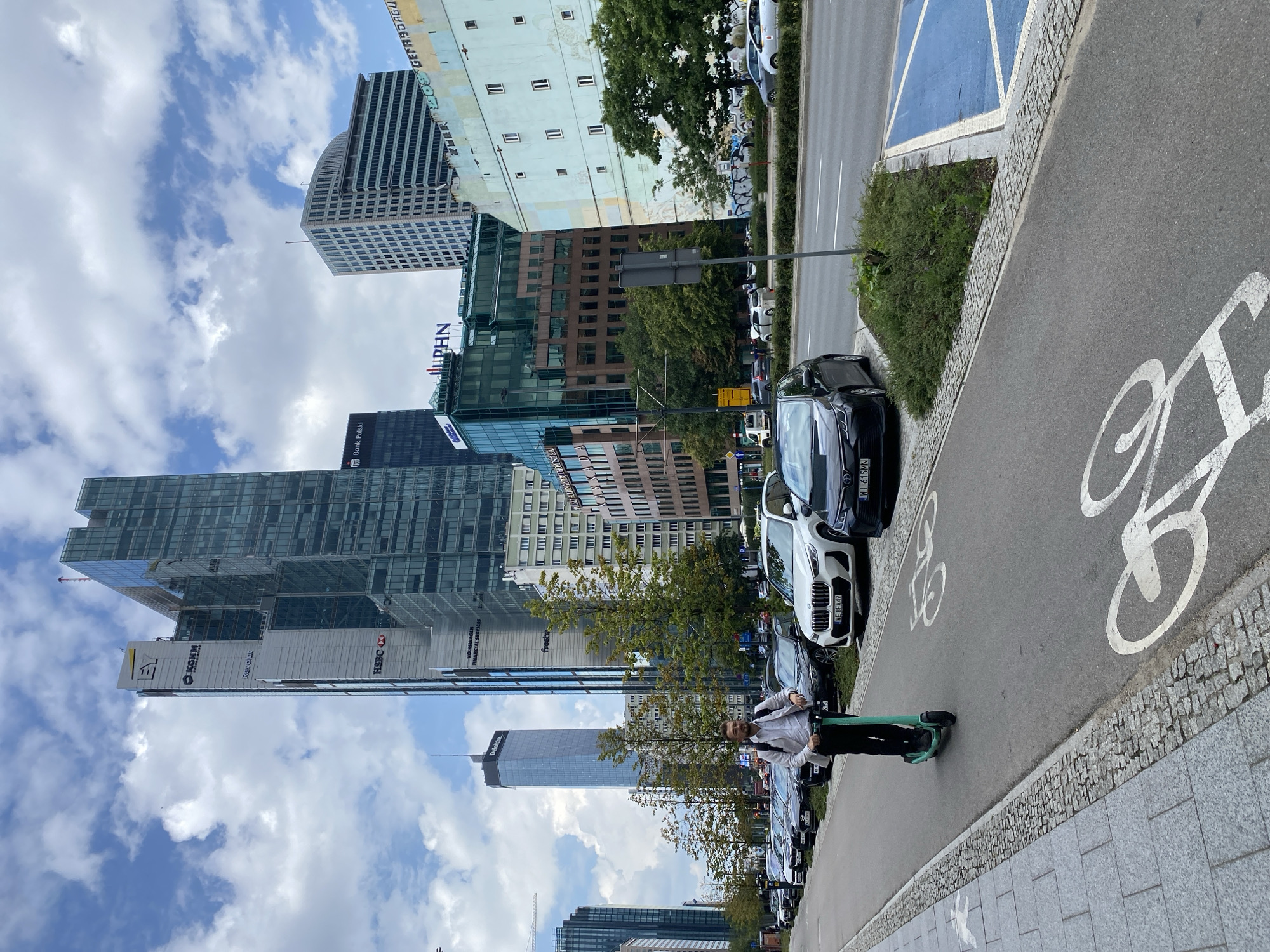 In Warsaw, Poland, on a cloudy day, a man rides an electric scooter on a bike lane bordered by a sidewalk and parked cars. Behind him, modern skyscrapers dominate the cityscape, including the EY, KGHM, and Bank Polski buildings, along with other office buildings and an older, pale blue building with graffiti. Cars are parked along the street, and trees line the sidewalk. The bike lane has white bicycle symbols painted on it.