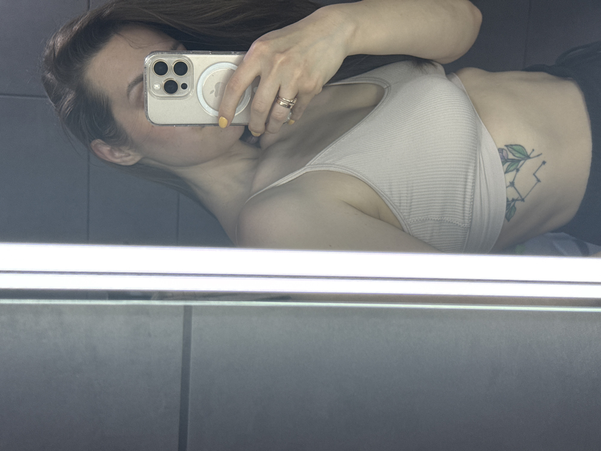 A mirror selfie depicts a female individual in an indoor setting within the city of Chaikovsky, Russia. The subject's reflection shows her upper torso, left arm, and the lower portion of her face, with long, dark hair partially obscuring her features. She is wearing a light-colored, ribbed spaghetti strap top. Dark lower garments are partially visible at the bottom of the frame. Her left hand, with fingernails painted bright yellow, holds an iPhone in a clear protective case, featuring a visible circular MagSafe accessory on its back, positioned to cover the upper part of her face. Two metallic rings are worn on the ring finger of her left hand. A tattoo, illustrating a star constellation connected to a purple flower with green leaves, is visible on her left side, below the ribcage. The background consists of dark grey walls, possibly tiled, with a prominent vertical strip of bright white light positioned to the right of the reflection.