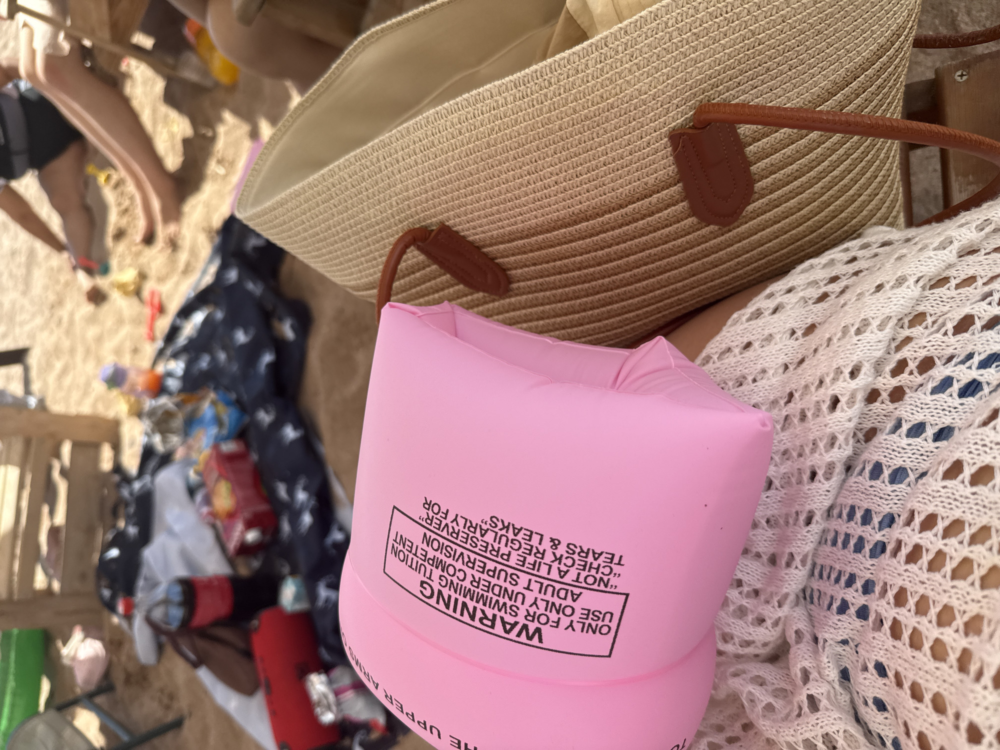 The image depicts a close-up of a beach scene in Sary-Oy, Kyrgyzstan. In the foreground, a pink inflatable arm float with black text warning against its use as a life preserver is held by a person wearing a white crocheted garment over blue shorts. To the right, a woven straw beach bag with brown leather straps is visible. The background shows a sandy beach scattered with various items, including a red suitcase, a bottle of Coca-Cola, and a dark blue patterned fabric. Several people are partially visible in the background, with their legs and torsos on the sand, suggesting they are relaxing or playing. The overall impression is a casual day at the beach, likely during warm weather, with children's toys also present on the sand.