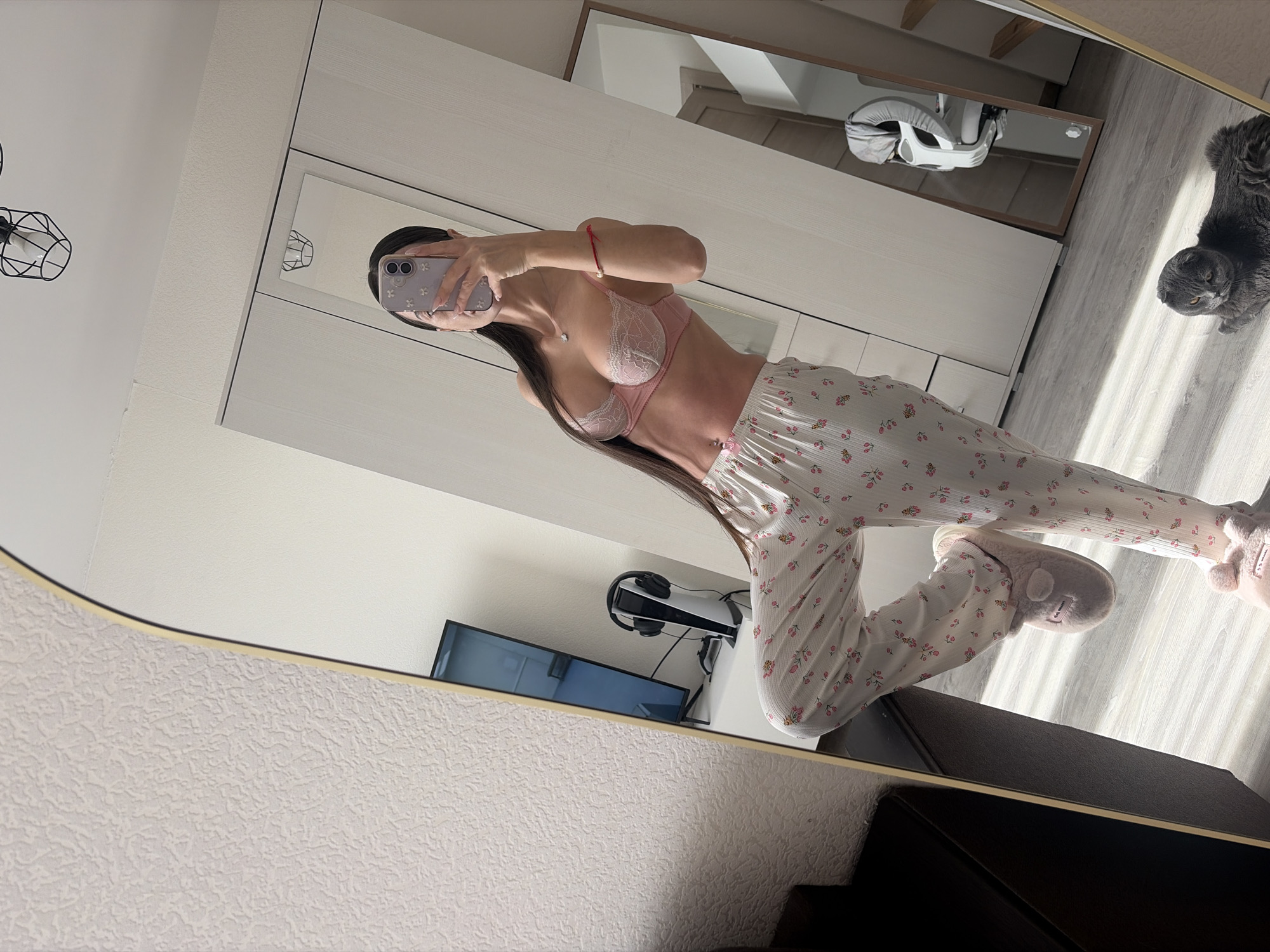 A mirror reflection reveals a woman in a light pink bra and floral pajamas, taking a selfie with a lavender-colored phone. Her long, dark hair flows down her back. The environment includes a gray cat sitting on the floor, a white baby swing, a white wardrobe, a television, headphones, and a PlayStation console. The room is filled with sunlight. There is no information on whether the location is in the city of Saint Petersburg, Russia.