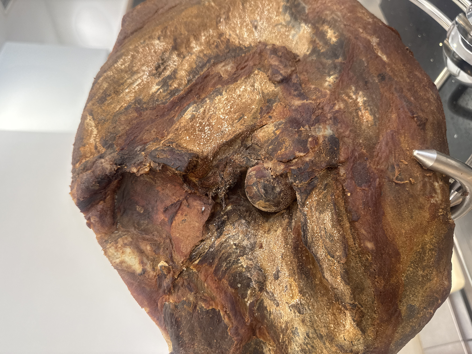 A close-up photograph shows a cured ham leg, likely Iberico or prosciutto, hanging from a metal hook. The ham has a mottled appearance, with shades of brown and gold, and textured, potentially mold-covered skin. The lighting suggests an indoor environment, and it could be in the city of Budva, Montenegro.