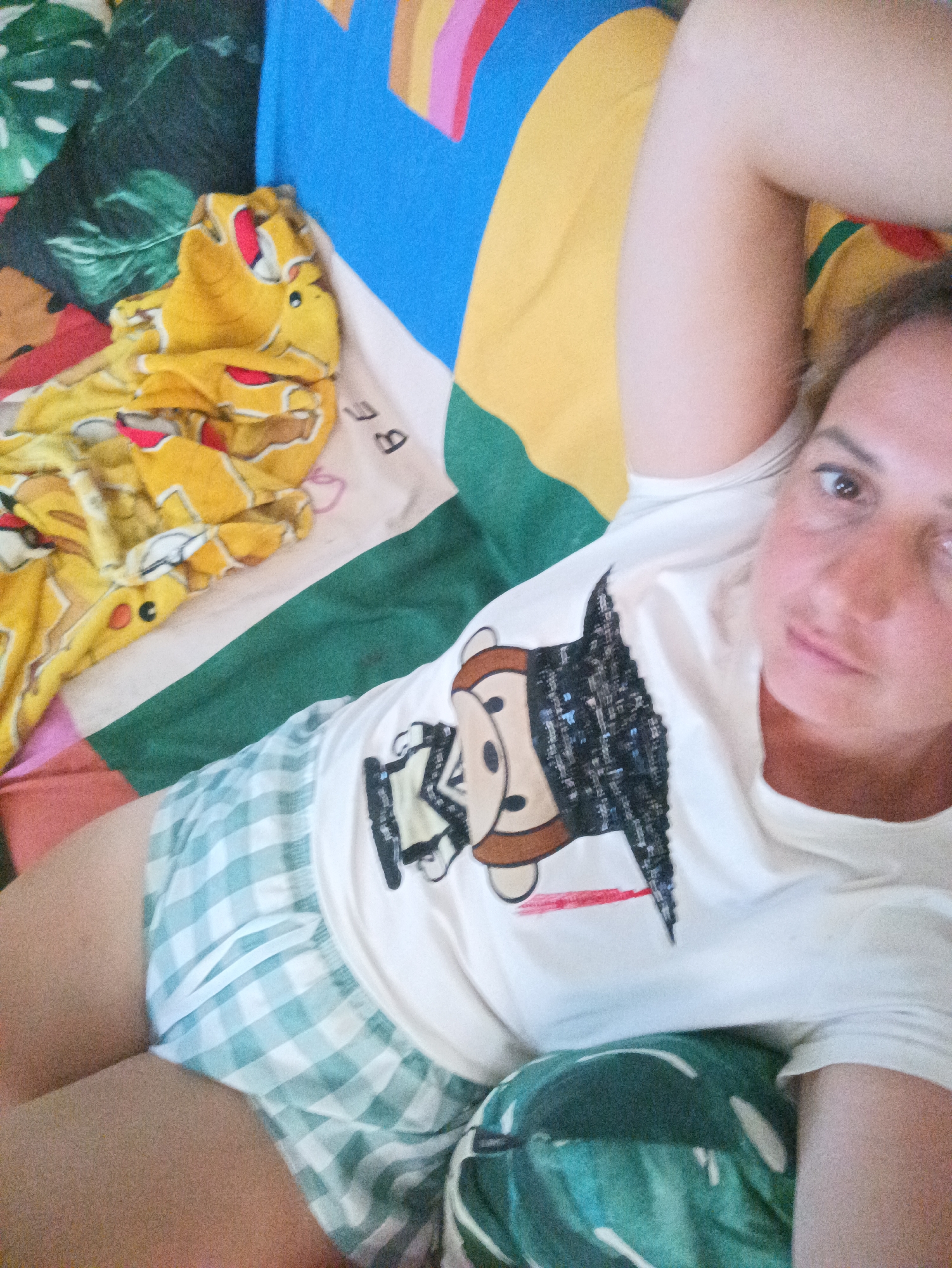 A woman lies on a bed in a room, possibly in Ban Nong Khon, Thailand. She wears a white t-shirt with a monkey design and green and white plaid shorts. The bed has a colorful patterned sheet and a yellow Pikachu-themed blanket. The environment appears domestic and informal.