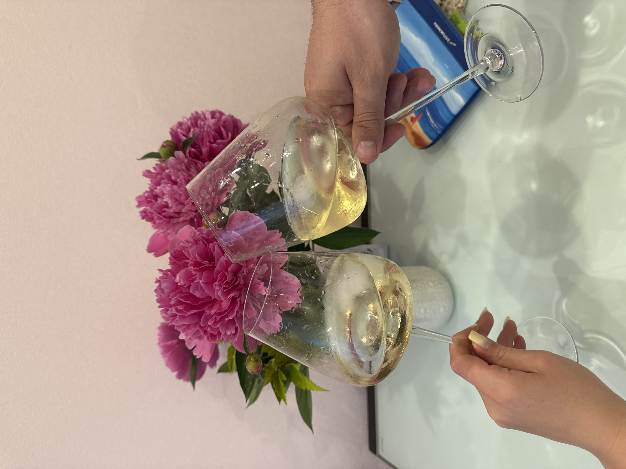 A high-angle, eye-level indoor photograph shows two individuals toasting with wine glasses over a light, reflective table. The scene is located in Vladimir, Russia.
In the background, against a plain light pink wall, a full bouquet of vibrant pink peonies with green foliage is displayed in a partially visible white, textured vase.
The foreground is dominated by two hands holding stemmed wine glasses. The left hand, appearing female with light skin and long, light yellow-tipped fingernails, holds a glass containing pale yellow liquid and visible ice cubes. The right hand, appearing male with tanned skin and shorter fingernails, holds a similar glass, also with pale yellow liquid and ice. Both glasses show condensation, indicating their cold temperature, and their bowls are clinking together in the act of a toast.
The reflective white table surface shows distinct reflections of the glasses, hands, and floral arrangement. To the far left, a dark rectangular object is partially visible. To the far right, a blue travel-related item, possibly a passport cover or brochure, is visible on the table, displaying a beach scene and the partially legible word "AEROFLOT".