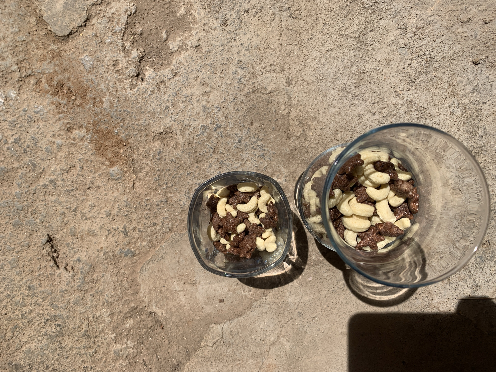 The scene, set in Rafin Par, Nigeria, features two clear glass containers placed on a rough, light-brown concrete surface under direct, strong sunlight. The larger container, a tall glass, is positioned in the upper right portion of the frame, partially cropped on its right side. It contains a substantial volume of mixed food items. Below and slightly to the left, a smaller, clear glass bowl holds a lesser quantity of the same food mixture. The mixture comprises light-colored, crescent-shaped cashew nuts and dark brown, irregularly shaped pieces, some distinctly star-like, resembling cereal or a crunchy snack. The concrete surface displays varying textures, minor cracks, and scattered debris. Harsh sunlight, originating from the upper left, casts sharp, distinct shadows of both containers, with the larger glass producing characteristic wavy shadow distortions. A prominent, dark, rectangular shadow is cast onto the upper right background, partially overlapping the shadow from the taller glass. No individuals or dynamic actions are present in the static composition.