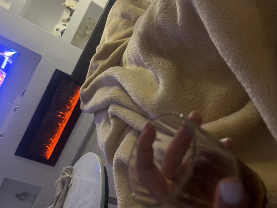 A person's hand holds a clear glass in front of a soft, beige blanket. An electric fireplace with a vertical orange glow is visible on the left side of the frame. A white, round table and a partially visible towel are near the fireplace. In the background, a television screen and decorative shelves are visible, completing the cozy indoor scene.