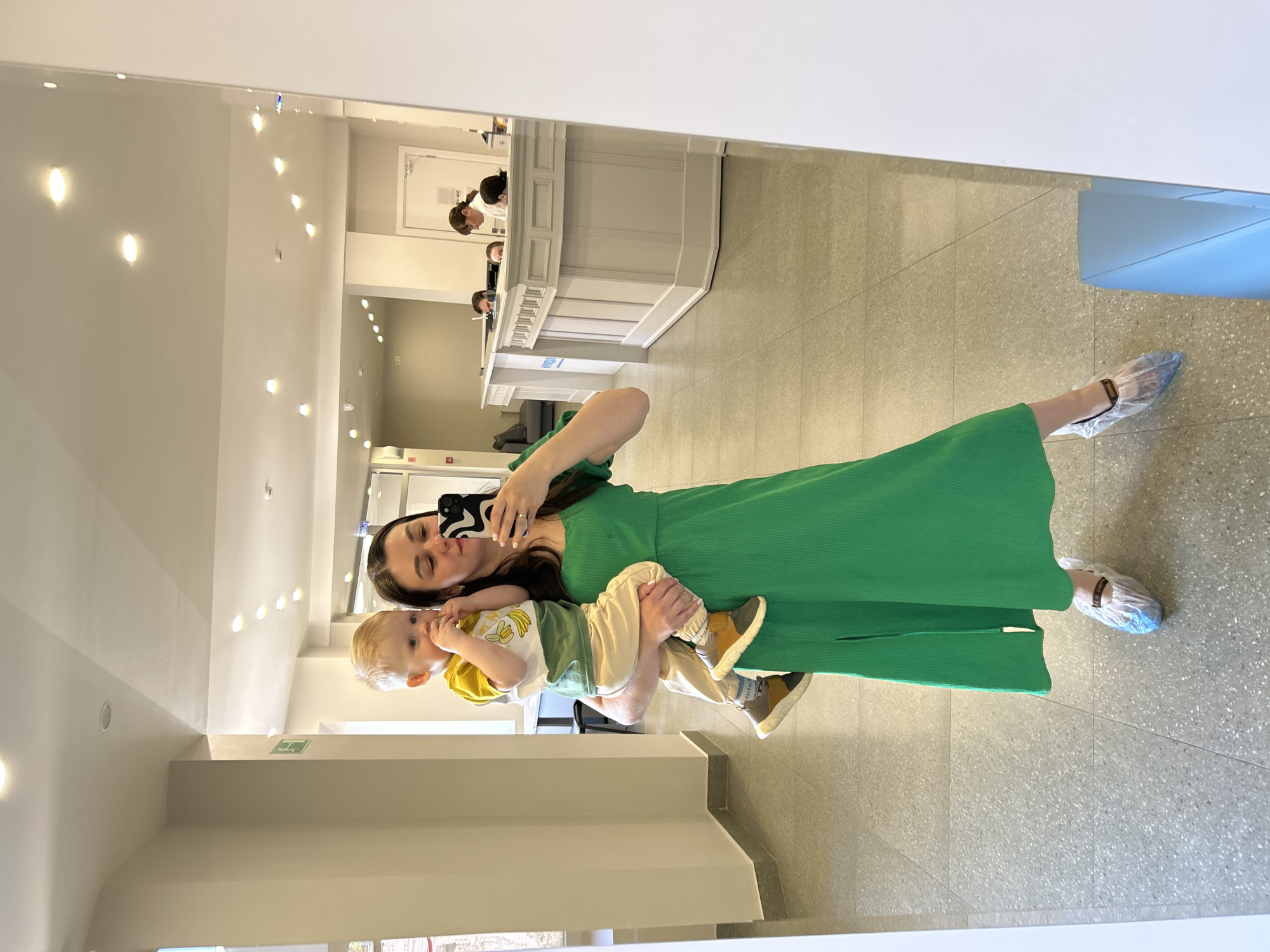 A woman in a green dress is taking a selfie in a mirror, holding a young child. The location appears to be a clinic or medical facility. The woman and child are wearing plastic shoe covers. A reception desk with personnel is visible in the background. The location is Izhevsk, Russia.