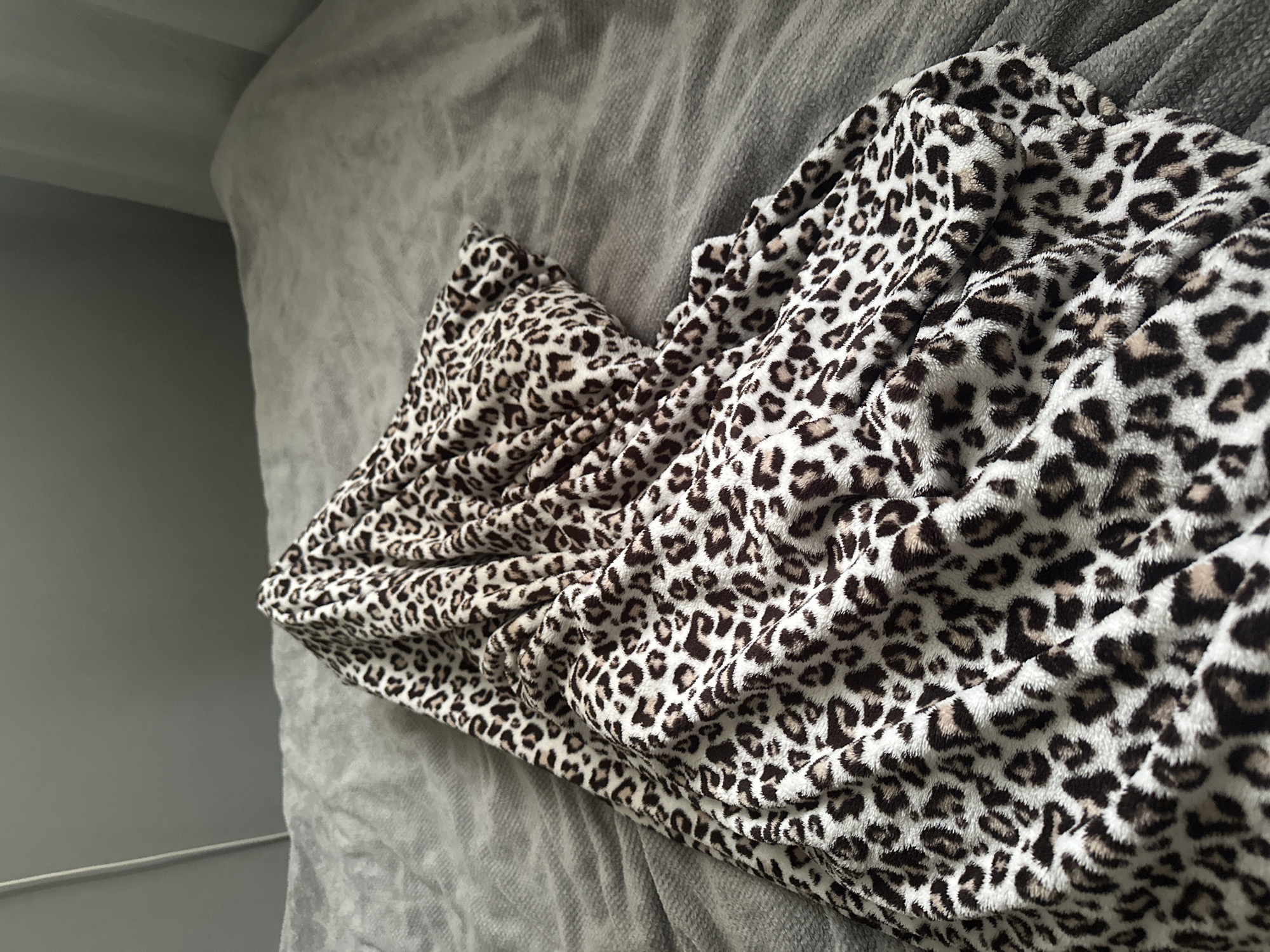The scene depicts an indoor environment, location: city of Unknown, Unknown. A textured, rumpled grey textile surface, resembling a blanket or duvet, occupies the majority of the frame. Resting upon this grey surface is a heavily folded and draped cream-colored textile featuring a distinct dark brown and tan leopard print pattern. The leopard print fabric is concentrated in the lower-left and central portions of the image, with its folds creating shadows that highlight its plush texture. In the upper background, a plain medium grey wall is visible, with a faint vertical light grey line on the far left, possibly a doorframe or wall edge. Towards the upper right, vertical folds of a lighter grey or white textile, likely curtains, are partially visible. No individuals are present in the image.