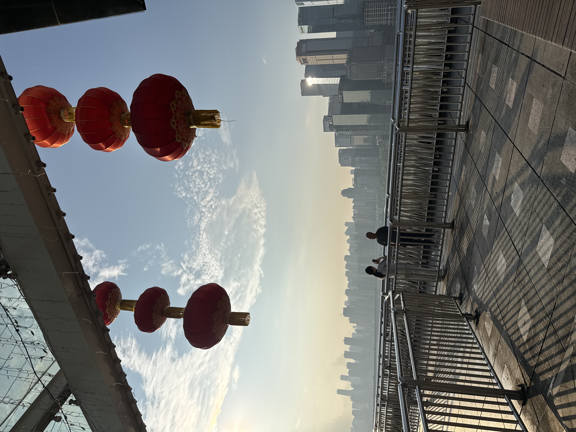 The image captures a view from a pedestrian bridge in Yuzhong District, China, overlooking a modern cityscape under a hazy sky. Suspended from the bridge's structure are several large, red, traditional Chinese lanterns. In the foreground, two individuals stand by a metal railing, one appearing to take a photo with a smartphone. The ground is paved with stone tiles, casting long shadows from the railing. The background reveals a dense collection of tall, contemporary buildings, indicative of a bustling urban center. The light suggests either early morning or late afternoon.