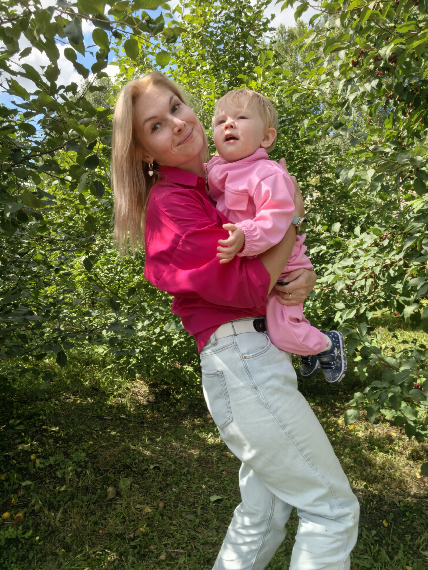 In Sterlitamak, Russia, a blonde woman wearing a vibrant fuchsia blouse and light blue jeans holds a baby dressed in a pink onesie. They are outdoors, surrounded by lush green trees and foliage, suggesting a park or garden setting. The woman is looking towards the camera with a slight smile, while the baby is looking upwards. The ground is covered in grass, with some fallen leaves and possibly fruit scattered around. The lighting indicates it is daytime.