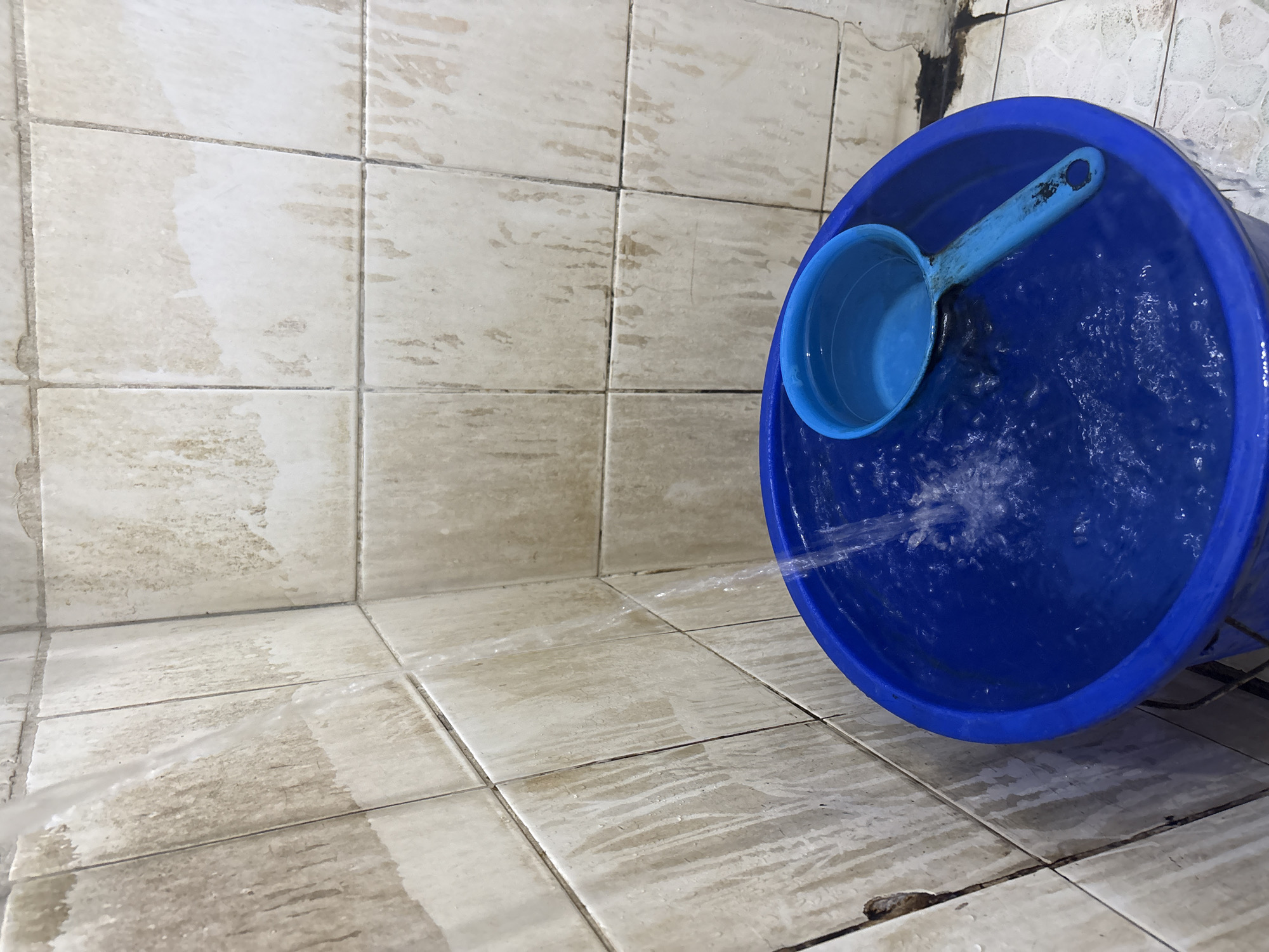 In a dimly lit bathroom in Palimanan, Indonesia, a blue plastic bucket overflows with water, a ladle resting inside. Water streams from an unseen source onto the surface of the water, creating ripples. The walls are tiled with pale, square tiles, stained with what appears to be mold or dirt.