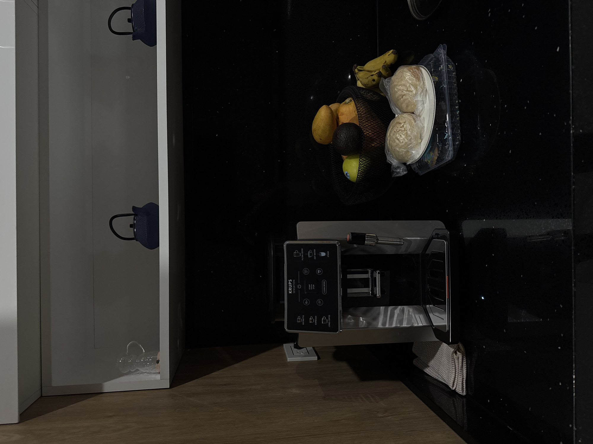 The image shows a kitchen scene. A white cabinet is on the left, with two blue teapots visible inside. On the countertop, there's a black wire basket containing fruit (bananas, a pear, an avocado, and a green apple). Next to the basket is a plastic container with two bread rolls inside. Adjacent to the fruit and bread, a Krups coffee machine is visible. A folded gray towel is located near the coffee machine. The countertop is black with white speckles, and the floor is light brown. There is no specific indication of the location being Puebla, Mexico.