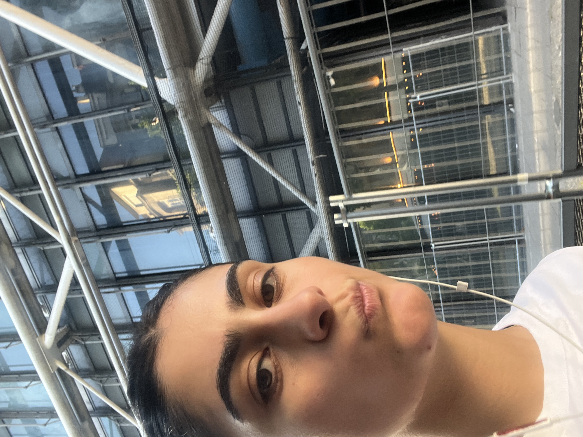 The image shows a woman taking a selfie in front of the Centre Pompidou in Paris, France. The building's distinctive exposed pipes and structural elements are visible in the background. The woman has dark hair and is wearing a white top. She is looking at the camera with a slight pout. An earphone wire is also visible. The building behind has a glass wall with light reflecting inside the rooms.