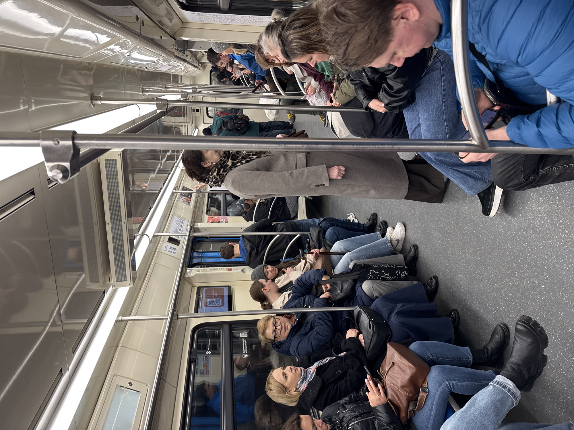The image shows the interior of a train, likely a subway car, packed with passengers. The car appears to be clean and well-lit with horizontal fluorescent lights. Most passengers are standing, likely due to the crowded conditions. The passengers are dressed in everyday winter clothes. Some passengers are looking at their phones. The train car's walls are a light color, and there are windows along the sides. The floor is gray. The train car's interior details suggest it is a relatively modern train. The presence of the Russian language in the image suggests it is located in Russia, and given the prompt's specification, likely in Khimki, Russia.