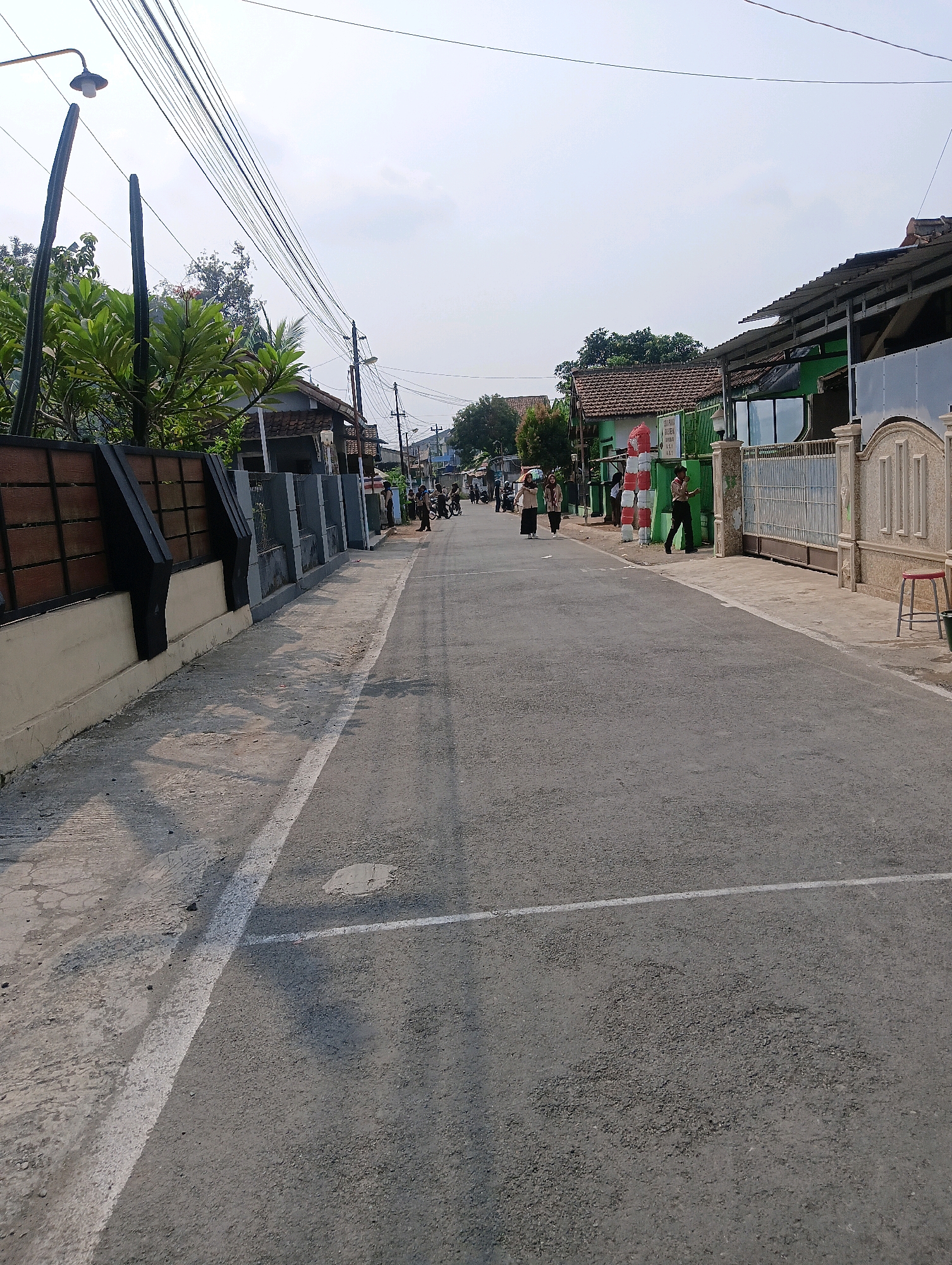 The image depicts a street scene in Sokaraja Wetan, Indonesia, under a clear sky. The street is paved with asphalt and marked with a white line. On the left side of the street, there is a concrete sidewalk with a wooden fence and some plants. On the right side, there are buildings, a metal gate, and a red and white striped pole. Several people are visible walking on the sidewalk and in the street, some carrying bags or riding bicycles. The buildings appear to be residential or commercial, with tiled roofs and green walls. Power lines are strung across the sky, supported by utility poles. The overall impression is of a typical residential street in an Indonesian town.