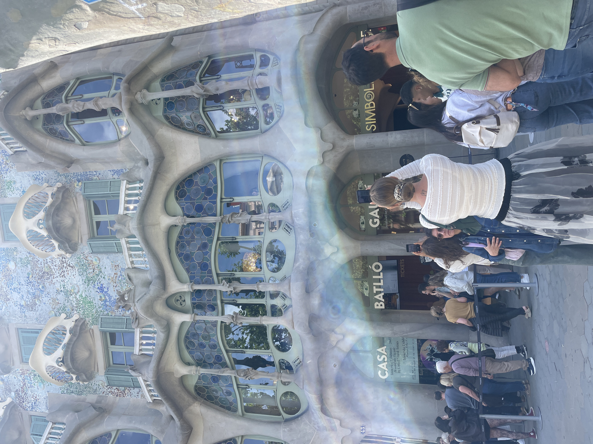 The image shows the Casa Batlló, a building in Barcelona, Spain. The foreground features a crowd of people, likely tourists, standing in a line. Some are taking pictures with their phones. The architecture is distinctive, with organic and whimsical shapes, especially around the windows and balconies. "Casa Batlló" is visible on a sign to the right. The word "tickets" is also visible on the sign.