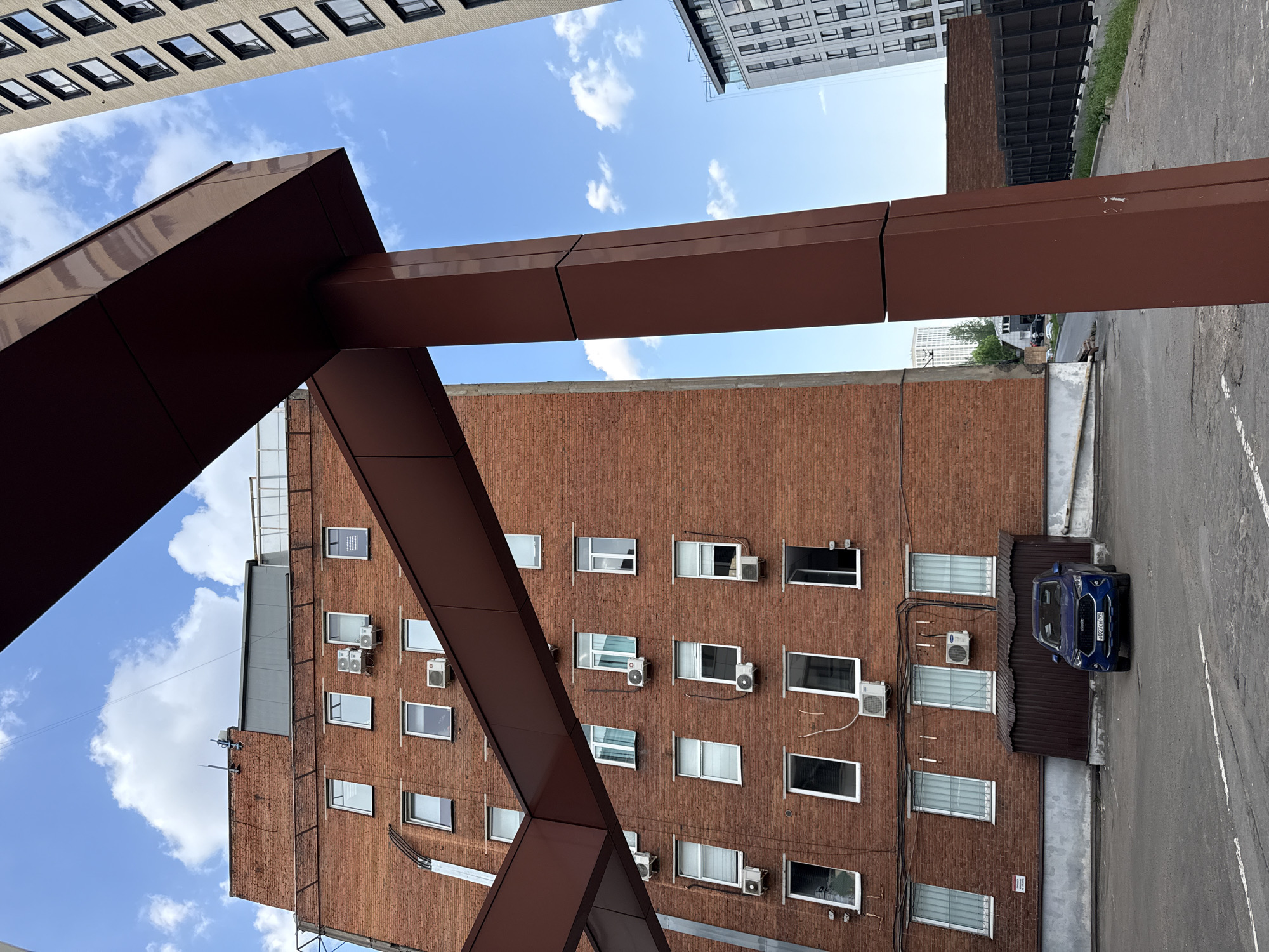 A street view in Khimki, Russia, presents an architectural juxtaposition of older brick structures and modern high-rise buildings. An orange-brick building with multiple windows, many fitted with air conditioning units, occupies the center. Beside it, a dark-blue vehicle is parked. Brown metal structures frame parts of the view, adding geometric shapes to the composition. Above, portions of taller buildings with repeating window patterns are visible. The sky is partly cloudy. The scene appears to be taken from a somewhat low angle, accentuating the height of the buildings.