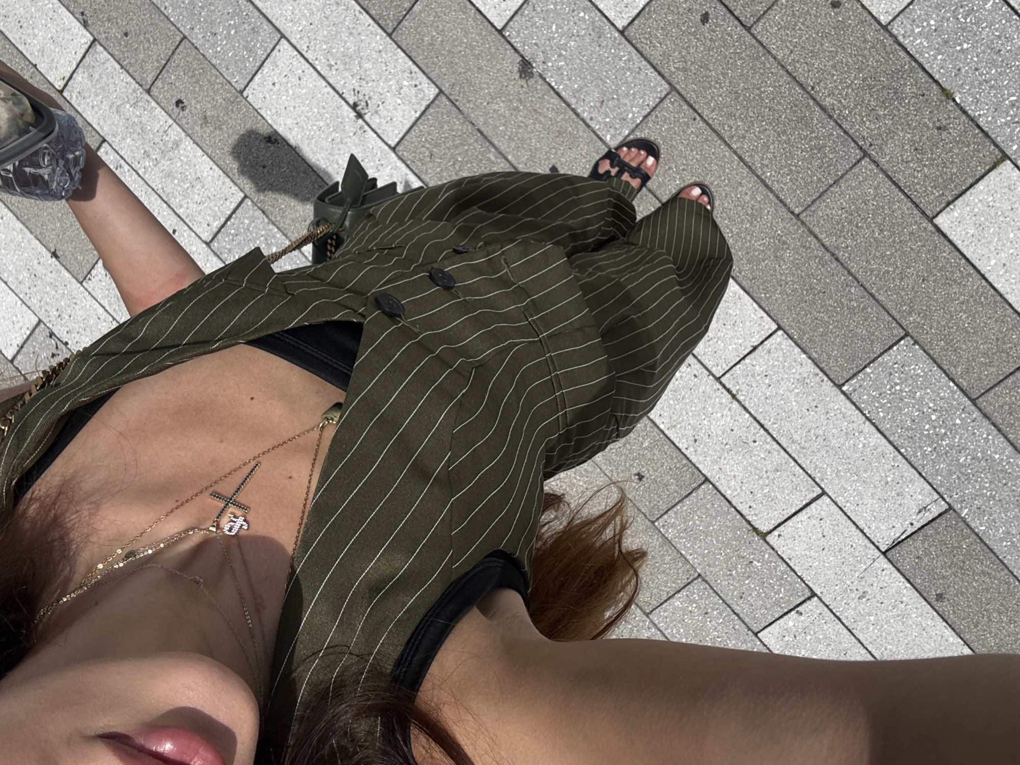 This media file is a close-up, overhead shot of a person standing on a tiled walkway. The subject is wearing a green pinstriped jacket over a black top, and black sandals. Two necklaces are visible on the subject's chest, one with a cross pendant. The setting appears to be outdoors, on a sunny day, given the strong shadows cast on the ground. The tiles are rectangular and arranged in a diagonal pattern, with varying shades of gray and white. The perspective of the shot is from above, looking down at the subject's body and feet, and also showing the ground. The subject's face is partially visible, with lips and part of the chin in focus. The overall tone of the image is casual and summery.