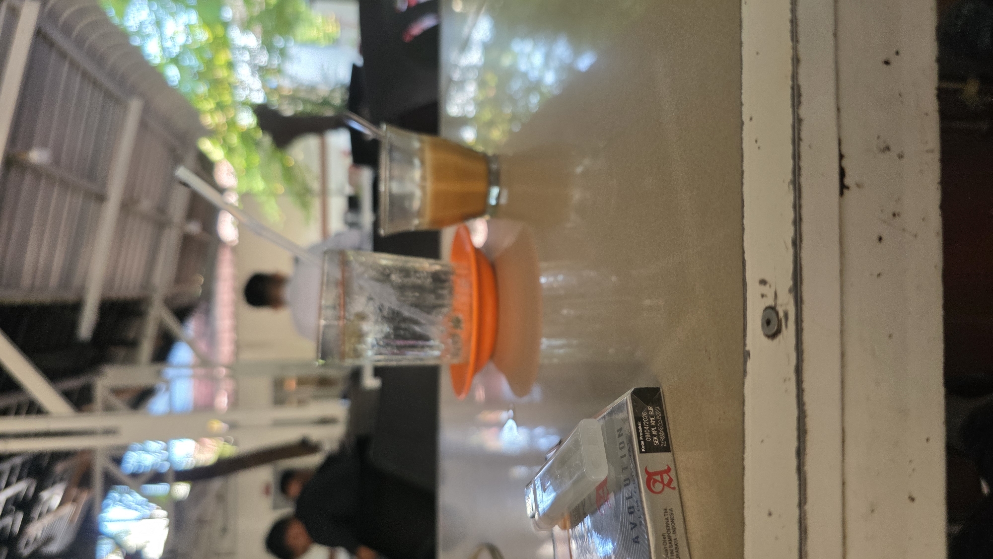 Here is a detailed description of the image:
**Scene Overview:**
The image appears to be taken at an outdoor cafe or restaurant in Lhokseumawe, Indonesia, as evidenced by the Indonesian text on the cigarette packaging. The focus is on a tabletop setting with drinks and cigarettes.
**Objects:**
* **Drinks:** There are two glasses on the table. One glass contains a light brown liquid, likely a coffee drink. The other contains a clear liquid, probably water. An orange saucer sits beneath the clear glass.
* **Cigarettes:** A pack of "AVOLUTION" cigarettes is visible, with Indonesian text indicating it is made by HM Sampoerna Tbk in Surabaya, Indonesia. The pack indicates a production code and date.
* **Table:** The table has a neutral colored surface (possibly beige or light gray). It is situated next to a white structure, which seems to be a support beam or a section of the building’s exterior.
**People:**
Out of focus, there are several individuals in the background. They are likely customers or staff at the cafe.
**Environment:**
The background is slightly blurred, indicating an open-air environment. There are structures suggesting a building exterior with an open-roof design that allows sunlight to filter through. There is a structure, possibly with a corrugated metal roof, visible towards the left.
**General Impression:**
The scene suggests a relaxed atmosphere at a cafe, common in Lhokseumawe, Indonesia. It captures a snapshot of everyday life with beverages and smoking materials on a table.
