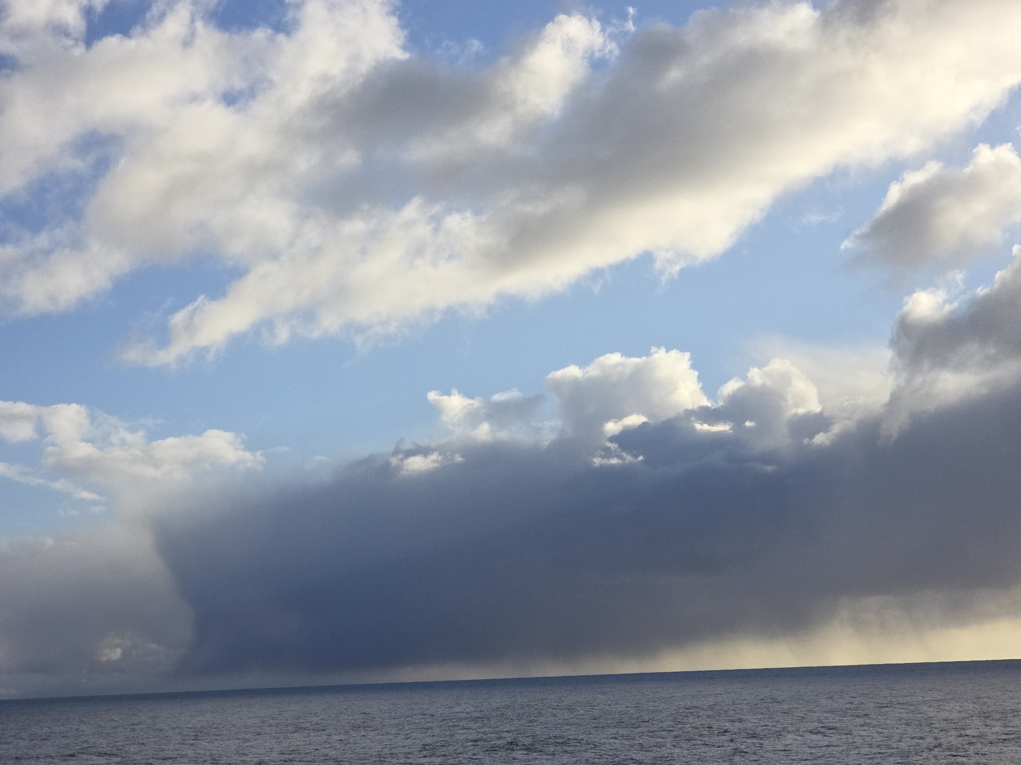 The image depicts a seascape with a distinct weather system in the distance. In the foreground, the calm surface of the ocean reflects the sky. Above the horizon, a large, dark rain cloud dominates a significant portion of the scene, with a visible curtain of rain falling from its base toward the water. Patches of clear blue sky and scattered white clouds are visible in the upper portion of the image, contrasting with the ominous rain cloud. The location appears to be open sea, far from any identifiable city.