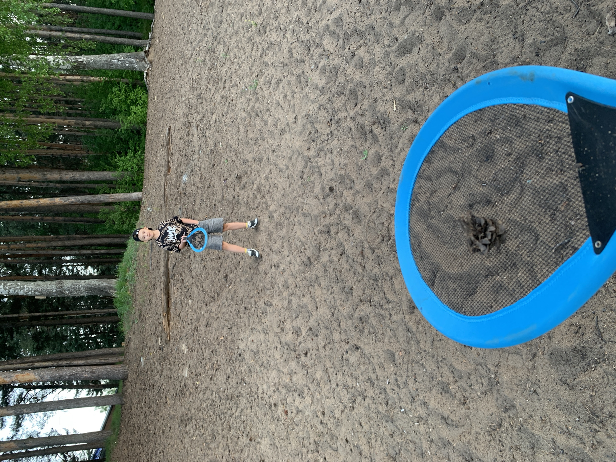 A boy stands on a sandy area, likely a playground or a park, holding a blue, round object. Part of the object is visible in the bottom right, and it appears to be holding a clump of debris. The background shows a line of trees. The location is identified as the city of Chaikovsky, Russia.