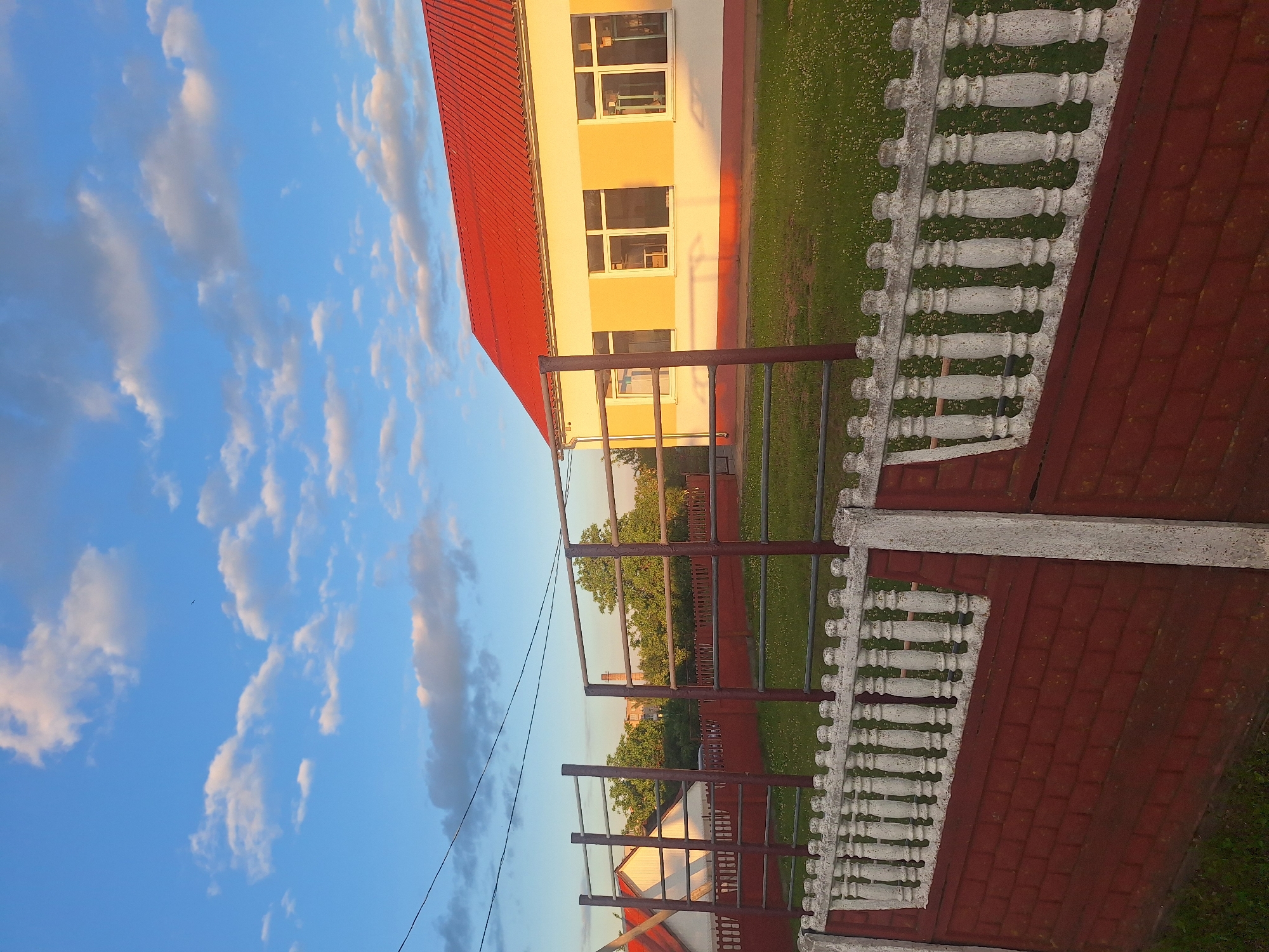 The image captures a scene at the edge of a schoolyard in the Капыльскі сельскі Савет, Belarus, during the golden hour. A red-tiled roof building with cream-colored walls and white-framed windows stands illuminated by the warm evening sun. In front of the building, a green lawn is dotted with small white flowers. A metal playground structure, consisting of horizontal bars supported by brown posts, is visible in the foreground, partially obscuring the building and the lawn. A brick and white concrete fence runs along the front of the property, separating the viewer from the schoolyard. The sky above is a clear blue, with scattered white clouds tinged with the colors of sunset. A thin black power line stretches across the upper portion of the frame. The overall impression is one of a peaceful, rural school setting as day transitions into evening.