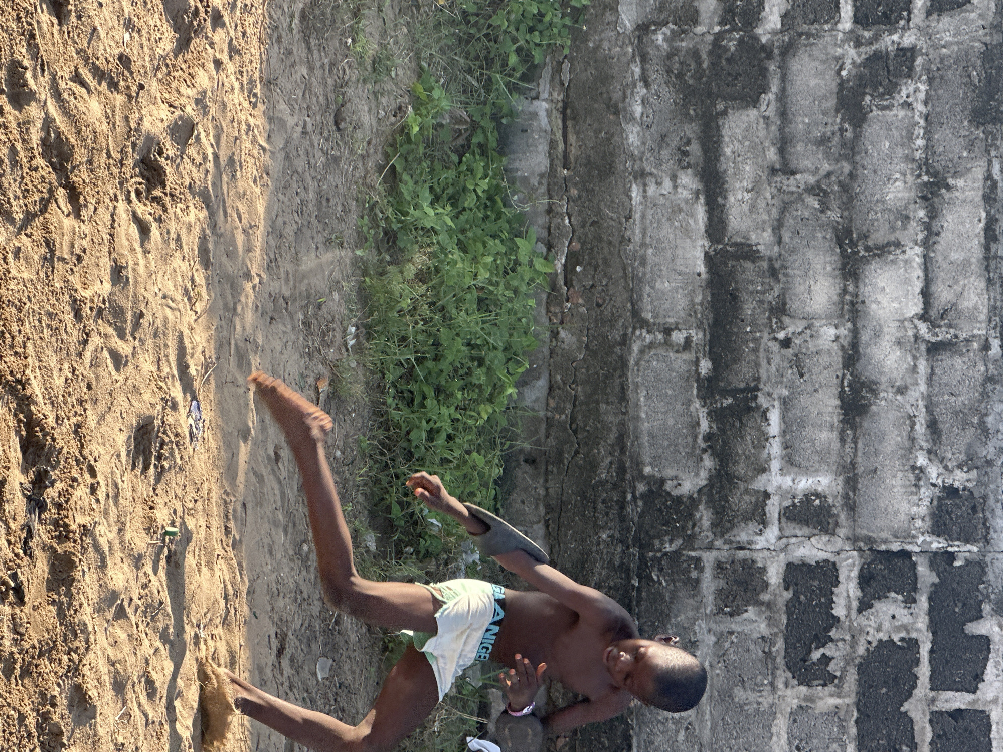 A young boy, appearing to be of African descent, is captured mid-stride in a sandy area in front of a rough, weathered brick wall. He is shirtless and wearing light-colored shorts with the word "NIGER" visible on the waistband. The boy's body is angled towards the right of the frame, with his right leg extended forward and his left leg bent, suggesting movement. He appears to be in motion, possibly running or kicking an unseen object. A dark, curved object is visible on his extended left arm. Green vegetation grows along the base of the brick wall. The ground is covered in dry sand, with indentations and tracks visible, indicating recent activity. The scene is lit by natural sunlight, casting shadows on the sand. The overall environment suggests an outdoor, informal setting within a city.