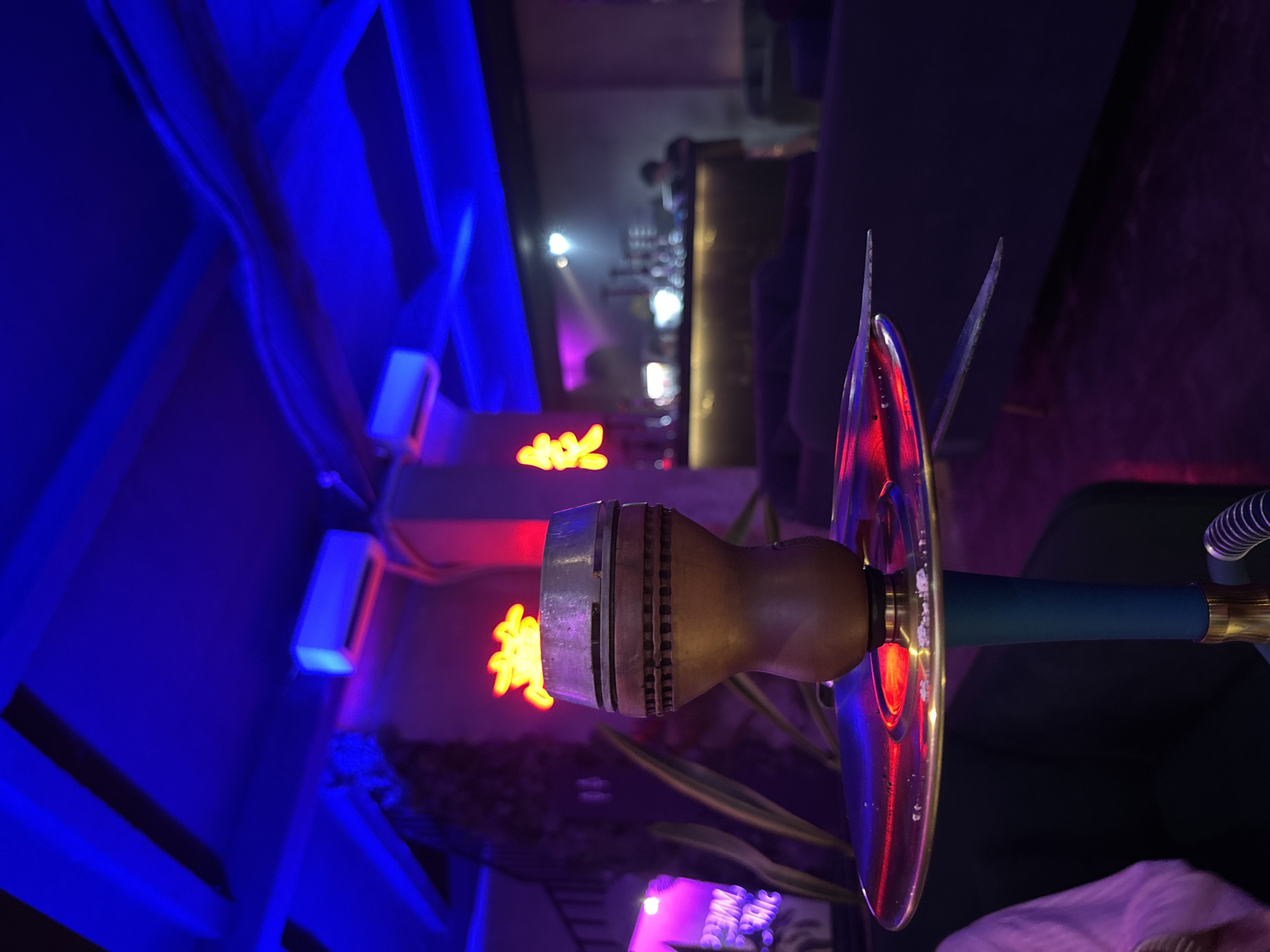 This image depicts an atmospheric indoor scene, likely a lounge or hookah bar during the evening or night. The main subject is a prominent hookah pipe, positioned on the right side of the frame. Its brass-colored bowl, detailed with ribs, sits atop a golden tray that vividly reflects surrounding red and blue neon lights. The hookah's stem is a dark teal.
The environment is characterized by deep blue and purple ambient lighting, creating a dim and moody atmosphere. On the left, vertical panels or curtains are intensely illuminated by bright blue light. Below these, two white rectangular fixtures on the wall also emit a blue glow. In the mid-ground, a neon sign displays two illuminated characters, likely Chinese, glowing in striking red and yellow and casting light onto the wall. The background is blurred, showing what appears to be a bar area with various bottles and scattered lights, suggesting a lively venue.
The scene exudes a chic, modern, and relaxed ambiance, typical of an establishment designed for socializing and unwinding. The artificial lighting, dominated by blues, purples, reds, and yellows, signifies a nocturnal setting. The only visible text consists of the red and yellow neon Chinese characters. The location context is Kuta Selatan, Indonesia.