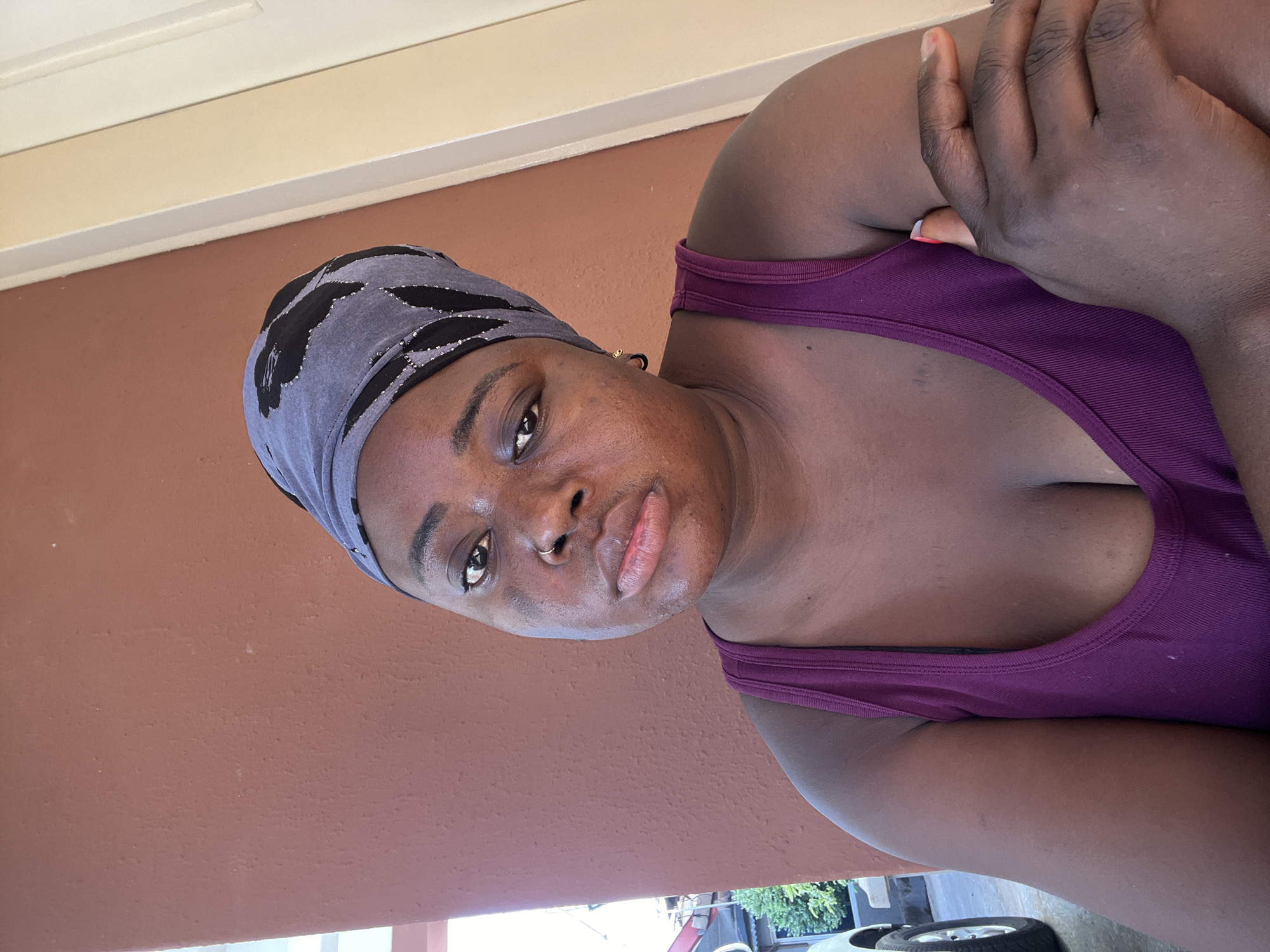 A dark-skinned woman wearing a purple tank top and a gray headscarf with black designs is seen indoors against a reddish-brown wall. She is positioned at an angle, with her hand raised near her chest. Outside the building, partial views of a car, plants, and tires are visible. The location is likely Jos, Nigeria.