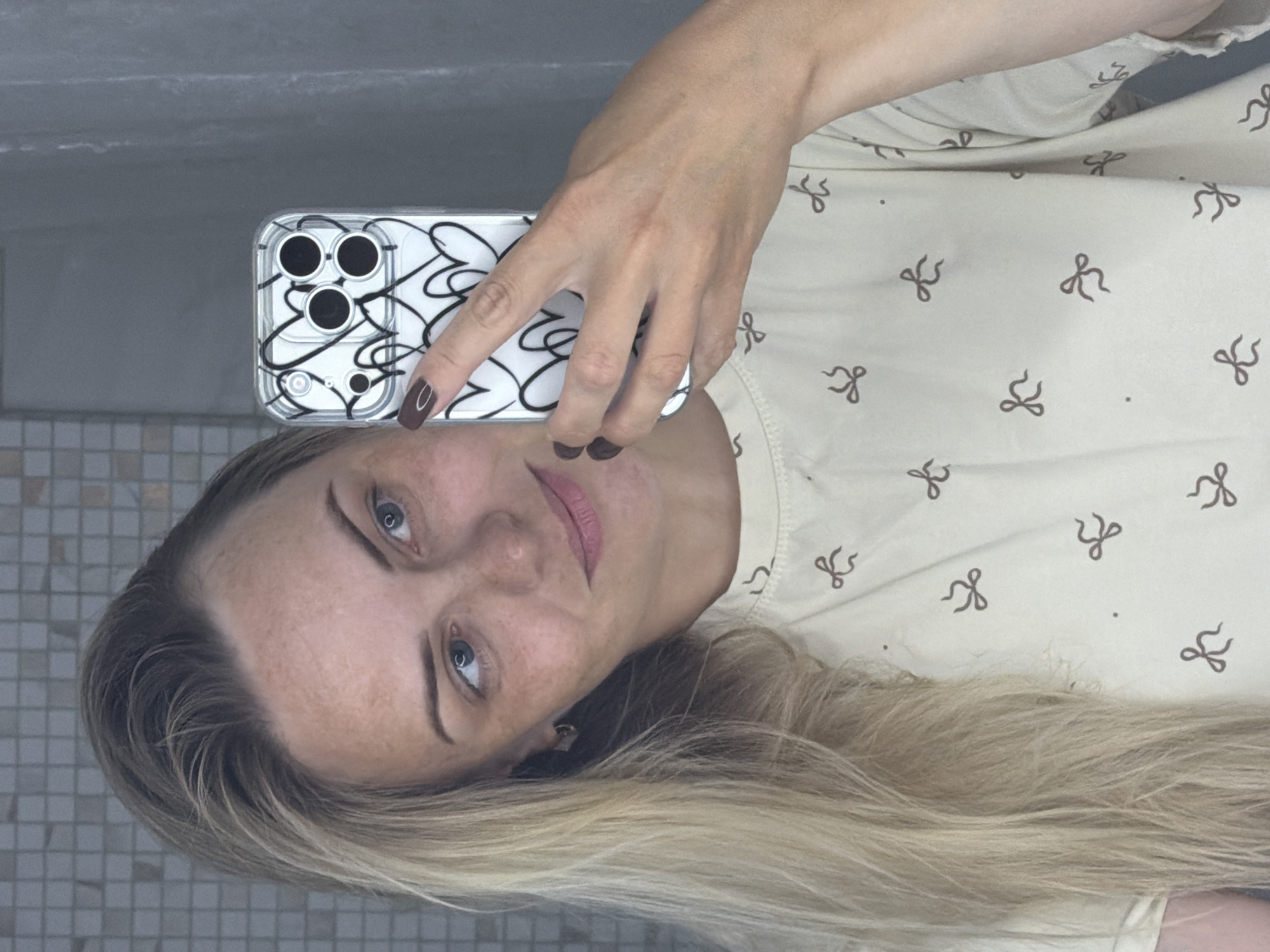 A young Caucasian woman with blonde hair and light blue eyes is taking a mirror selfie in what appears to be a bathroom. She is wearing a light-colored t-shirt with a pattern of small brown bows. She is holding a white smartphone with a clear case featuring a black heart design in her right hand, partially obscuring her face. Her left arm is visible. The background shows a tiled wall and a grey shower curtain. There are no discernible landmarks or environmental features that would indicate a specific location in Votkinsk, Russia. The image captures a personal moment of the woman.