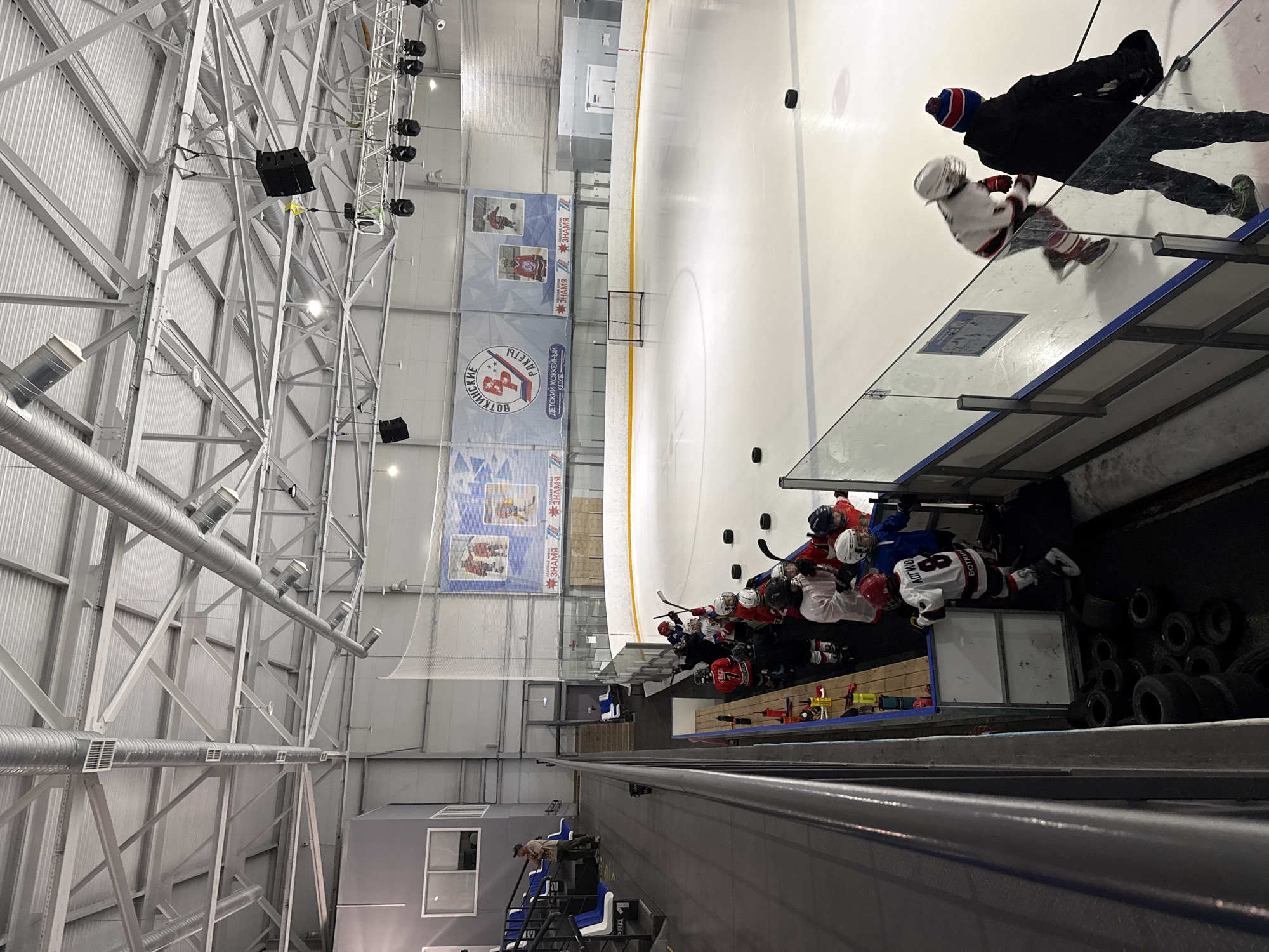 This image depicts an indoor ice hockey arena, capturing a children's hockey practice or training session. The viewpoint is elevated, offering a perspective down onto the ice rink and the surrounding areas.
On the bright white ice surface, a small practice goal is visible, along with several black hockey pucks. Near the boards on the right, a group of young hockey players, dressed in full gear including helmets, colored jerseys (red, white, blue), and skates, are gathered around the player bench. Some players are seated on the bench, while others stand holding their hockey sticks. One player's white jersey features the number "88" and "BOTK NHC" (likely "Votkinsk Hockey Club" abbreviated), and another player wears number "7". An adult, possibly a coach, wearing a black jacket and a red and blue beanie, stands on the ice beside one of the young players, who is also on the ice near the boards.
The arena's infrastructure is prominent, with a robust metal truss system supporting the high ceiling. Large silver ventilation ducts run across the upper part of the space, accompanied by suspended speakers and artificial lighting fixtures. Along the back wall, several colorful banners are displayed. Visible Russian text on these banners includes "ВОТКИНСКИЕ РАКЕТЫ" (Votkinsk Rockets), "ДЕТСКИЙ ХОККЕЙНЫЙ КЛУБ" (Children's Hockey Club), and the repeated word "ЗНАМЯ" (Banner/Flag), clearly identifying the club and its location (Votkinsk, Russia). Below the viewing area on the left, a person is seen near some spectator seating. A stack of black tires, commonly used in hockey training, is visible behind the player benches. The scene is well-lit by artificial lights, indicating an active training environment.