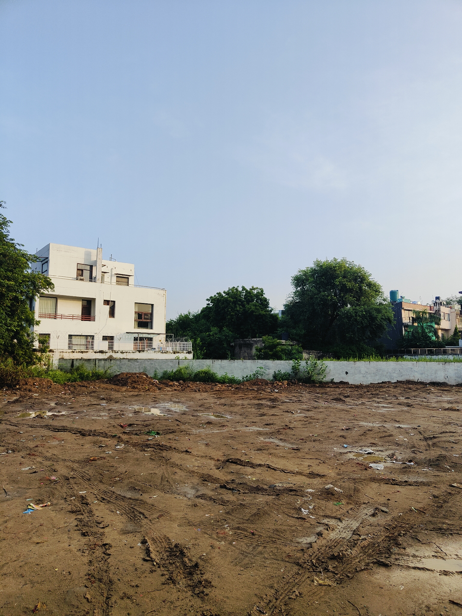 This is a ground-level, wide-angle photograph taken outdoors in Gurgaon, India. The scene depicts a construction site or undeveloped plot of land. The foreground is dominated by uneven, muddy ground with visible tire tracks from vehicles, indicating recent activity or disturbance. Scattered debris, including plastic and paper, is present on the ground. Puddles of water are also visible in the muddy area, suggesting recent rain.
In the mid-ground, a low, light-colored concrete wall runs horizontally across the image, separating the muddy foreground from the area behind it. Beyond the wall, several buildings and lush green trees are visible. On the left, a modern, white, multi-story building with multiple windows and balconies stands prominently. The building has a flat roof and appears to be an apartment complex or office building. To the right of this building, dense green trees form a thick canopy. Further to the right, other buildings with varying architectural styles and colors, including some with visible water tanks on their roofs, are nestled among more trees. The sky above is a clear, pale blue with very light wispy clouds. The overall impression is a transitional urban landscape, possibly on the outskirts of a developed area, with evidence of construction or demolition and natural elements.