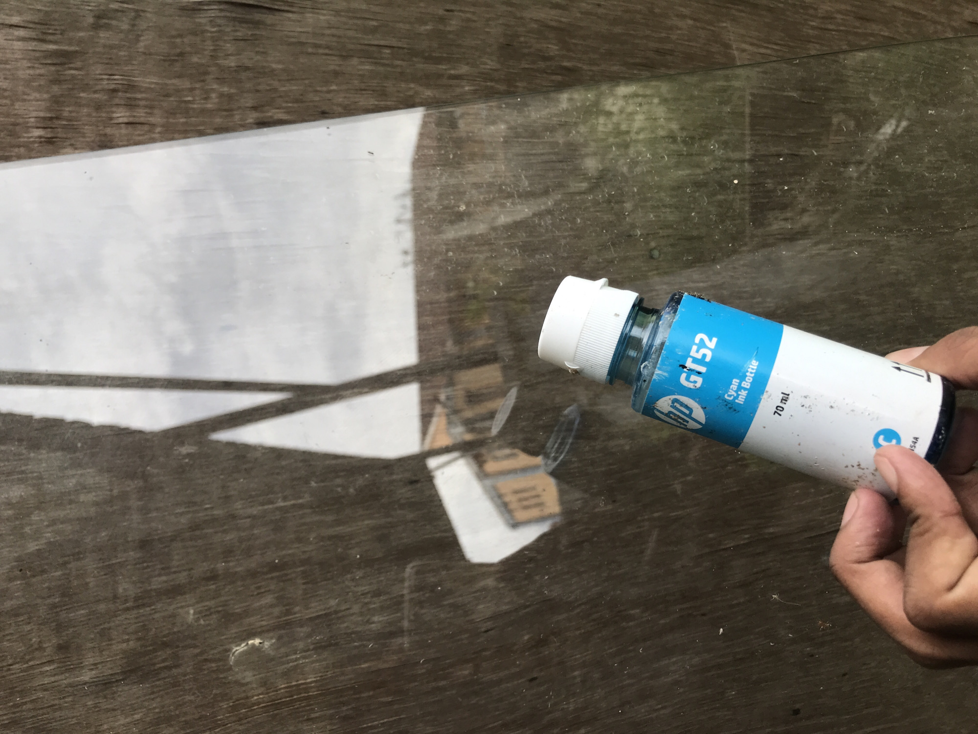 The image shows a hand holding an HP GT52 cyan ink bottle over a reflective glass surface resting on weathered wood. The glass reflects a building or object with sharp angles, potentially part of the urban landscape. The background appears to be part of the city of Lhokseumawe, Indonesia, indicated by the weathered wood and the style of the surrounding reflections. The image doesn't capture any direct action or interaction; it focuses on the positioning of the ink bottle and its reflection within the setting.