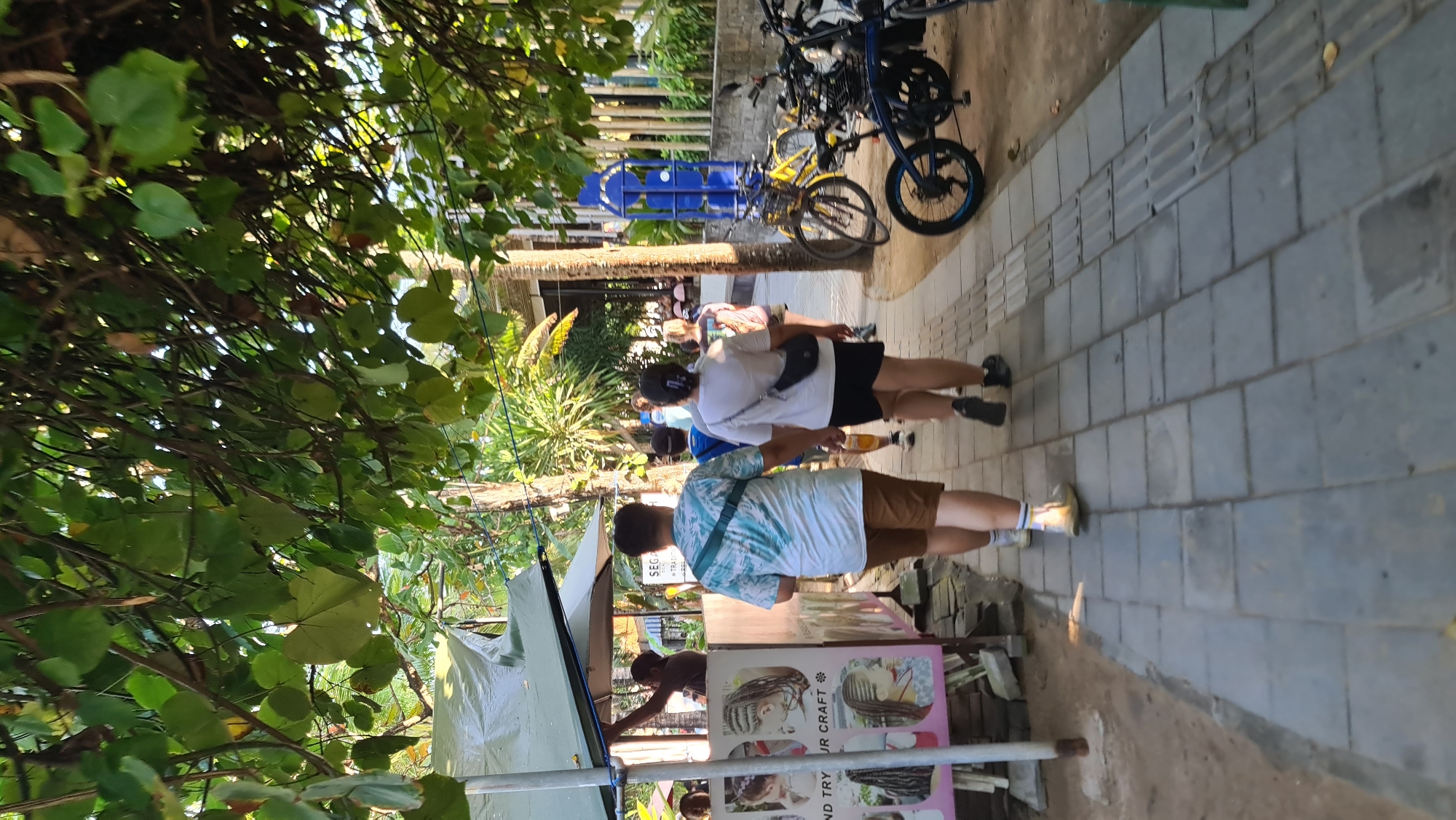 In Denpasar, Indonesia, a paved walkway lined with trees and tropical foliage leads past a temporary stall advertising hair braiding services. A man in a patterned blue and white shirt and brown shorts walks away from the camera, his arm extended. Beside him, a woman in a white t-shirt and black shorts with a black crossbody bag also walks away. Several other individuals are visible further down the path, including someone in a blue shirt and another in a pink t-shirt. In the background, bicycles are parked, and a bright blue, multi-panel sign stands vertically. The scene appears to be a public area, possibly a street or a path near a beach, with evidence of commerce and pedestrian activity.