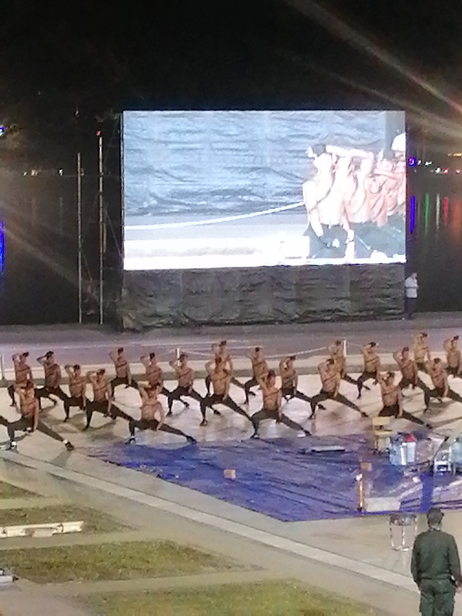 A group of men, appearing to be dancers or performers, are positioned on an outdoor stage in front of a large LED screen. They are wearing dark bottoms and are bare-chested, with many in athletic poses, such as lunges with one arm raised. The LED screen displays an image of other shirtless individuals, possibly a pre-recorded performance or a visual element for the current show. The scene takes place at night, indicated by the dark sky and stage lighting. The background shows some reflective water, suggesting the performance might be near a lake or river. A single person in a dark uniform is visible walking away from the stage in the lower right corner of the frame. The overall setting is a public outdoor venue, possibly a festival or concert, with a large audience implied to be present, although not directly visible in the frame. The time of day is clearly nighttime. There is no visible text. The activity is a performance or dance routine.