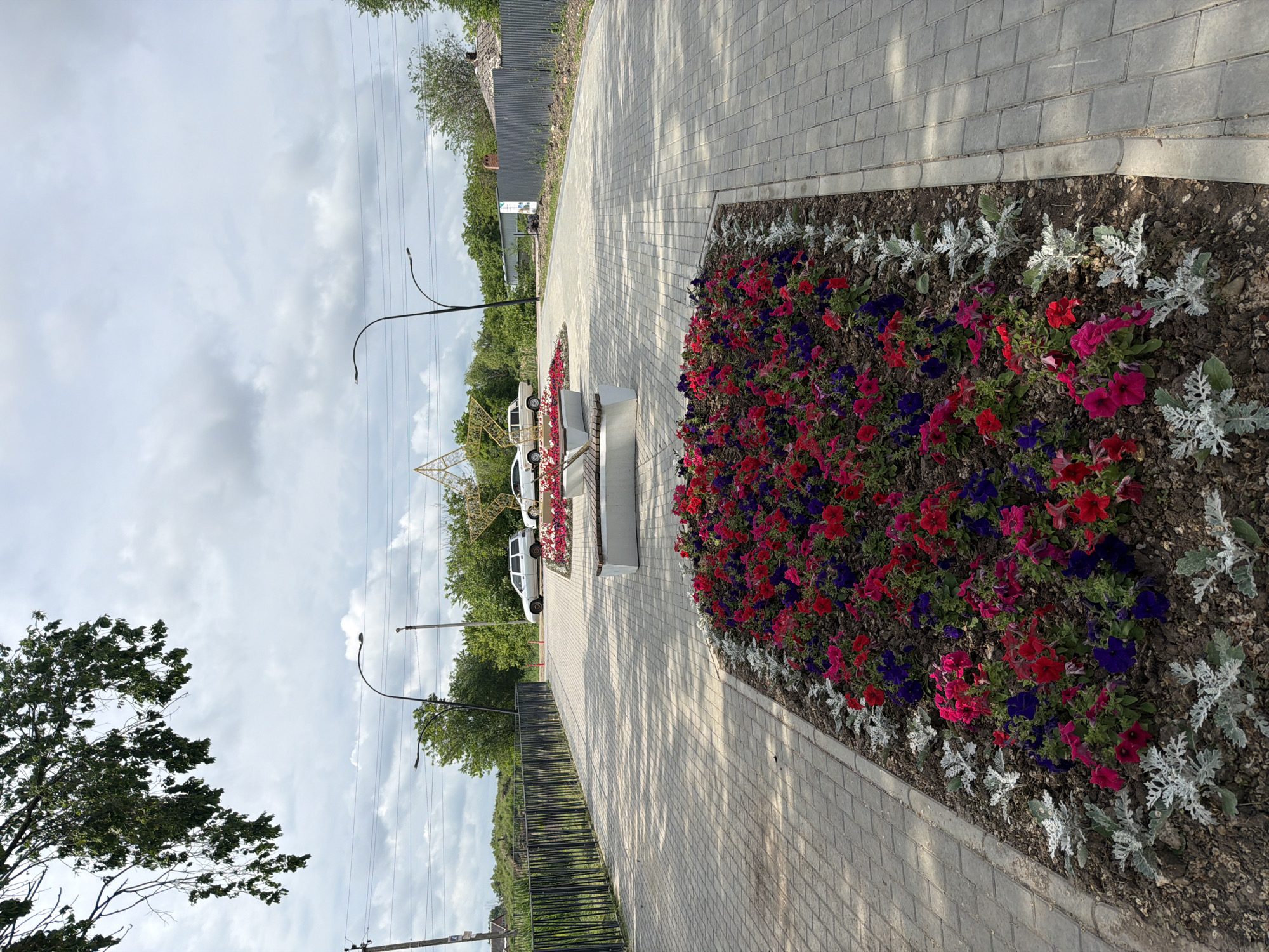 The image shows a paved walkway in the city of Leningradskaya, Russia. On the right side of the walkway is a flowerbed with red, pink, and purple flowers bordered by silver-leafed plants. The walkway leads to a white bench. Beyond the bench, there are parked white vans and green trees. A metal fence runs along the left side of the walkway. There are lampposts on either side of the walkway. The sky is cloudy.