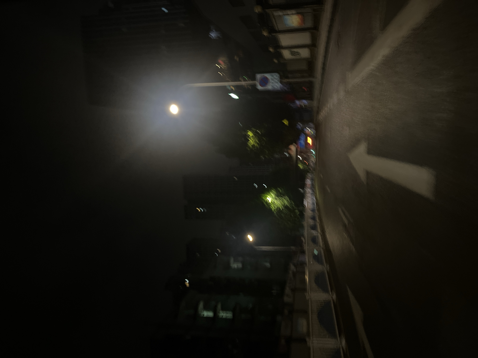 The image displays a blurry, dark, nighttime scene of an urban street in Yuzhong District, China, viewed from a low perspective, likely from a moving vehicle. The wet or reflective asphalt road surface is prominent in the foreground and midground, featuring a large, white straight-ahead arrow painted centrally, with a fainter white line to its right.
Along the left side of the road, a white barrier or fence with dark, arched recesses runs parallel to the street. Behind this barrier, dark, multi-story buildings are visible, some exhibiting indistinct illuminated windows or internal lights. Several streetlights on the left emit a soft, yellowish-orange glow.
On the right side of the road, a tall street light pole stands prominently, emitting a bright, diffused white light that creates a substantial lens flare in the upper right quadrant of the image. A circular white and blue sign is attached to this pole, illegible due to blur. Further to the right, dark structures, possibly walls or fences, are visible, interspersed with several rectangular, subtly illuminated panels.
The background consists of dark, indistinct silhouettes of tall buildings against a dark sky. Scattered throughout the scene are numerous small, blurry light sources, appearing as streaks or crescent shapes, indicative of distant city lights and motion blur. No people or clearly identifiable vehicles are discernible, though distant red and yellow lights on the road ahead suggest other traffic.