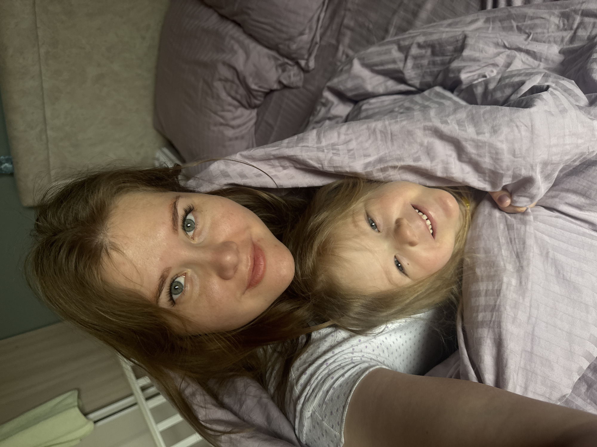 The image shows a woman and a child lying in bed under a purple striped comforter. The woman has light skin, auburn hair, and blue eyes. The child has light skin, blonde hair, and blue eyes. Both are smiling. They are indoors, likely in a bedroom. The location may be Gornyi (Dvurechenskoe S/P), Russia.