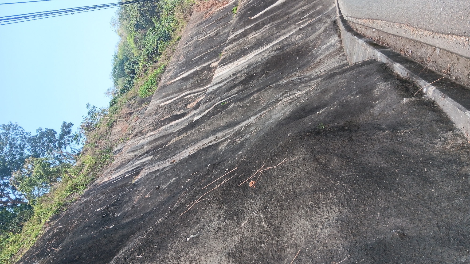 The image depicts a steep, dark gray rock face, possibly a cut for a road. The rock is uneven and has streaks of lighter gray and white running diagonally down its surface. Vegetation, including trees and shrubs, grows at the top of the rock face and further up the slope, with the sky visible above. In the foreground, a paved road curves to the right, with a concrete curb and drainage ditch running along the base of the rock face. There are no people or animals visible in the image. The overall scene suggests a natural landscape alongside a man-made road, likely in a tropical or subtropical region given the lush vegetation and clear sky.