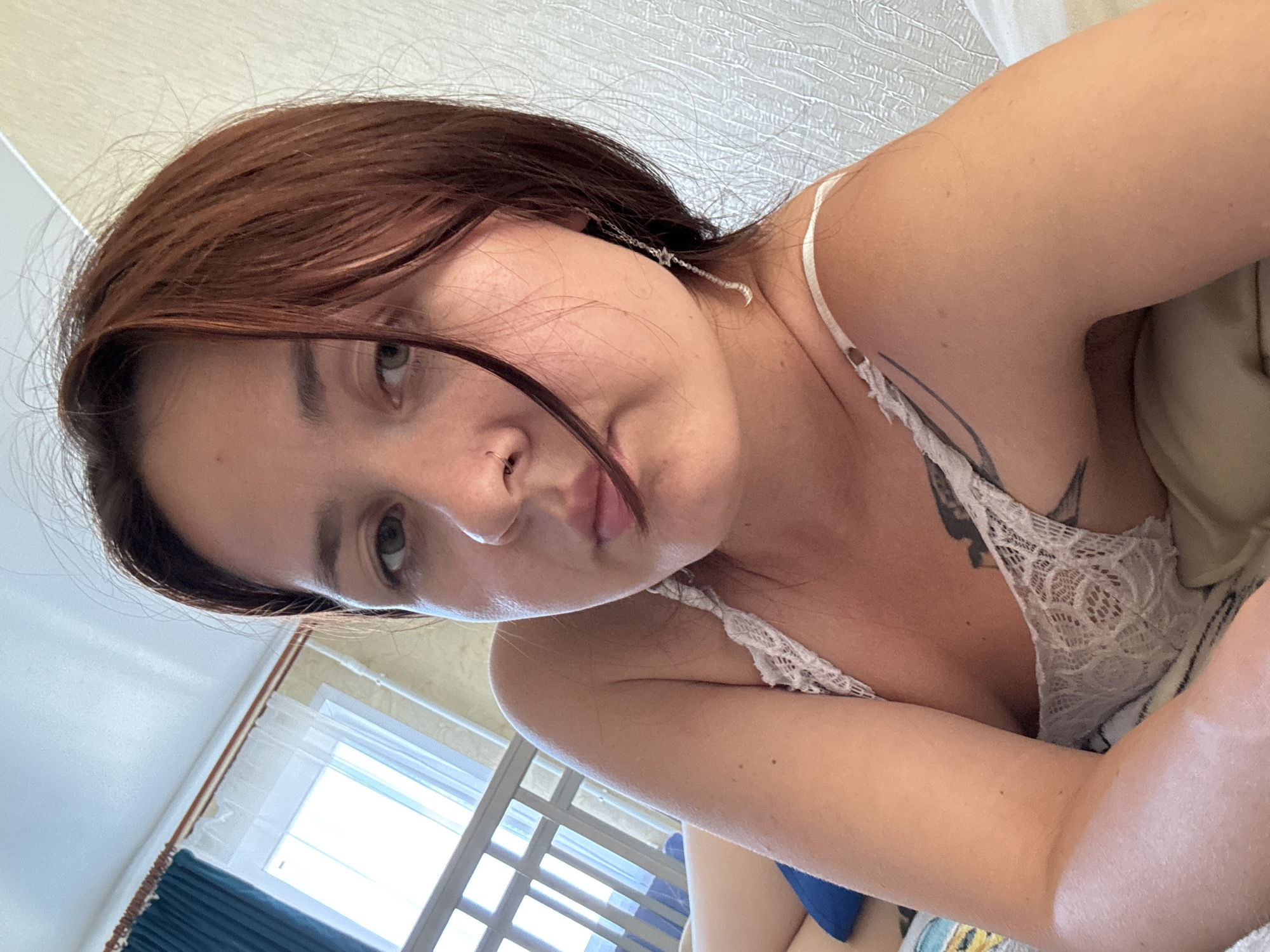 A young woman with auburn hair, wearing a white lace bralette, is lying on a bed in a room. She is looking directly at the camera with a neutral expression. Her left arm is bent and resting on a pillow. A tattoo is visible on her upper chest. Behind her, a window with blue curtains and sheer white blinds is seen. A crib or a similar structure with bars is partially visible to her left. The room appears to be a bedroom, possibly in an apartment. The lighting suggests it is daytime.